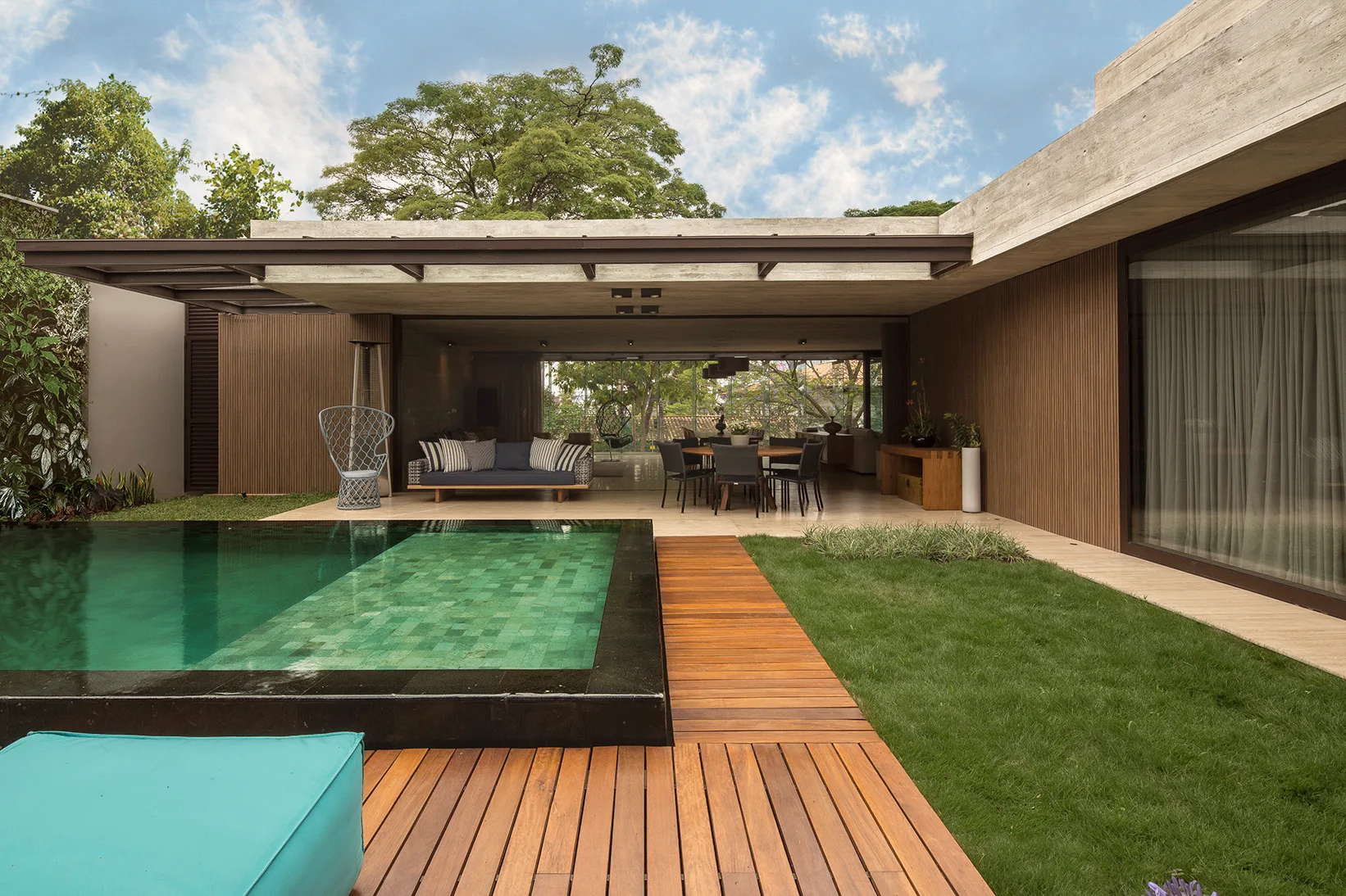 A rectangular swimming pool is flanked by a wooden deck and a grassy lawn next to a covered modern terrace with slatted wood paneling.