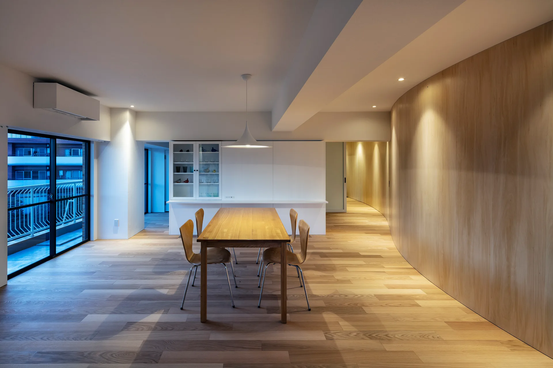 A minimalist dining room features a wooden table and chairs set against a large curved wood-paneled wall under a white pendant light.