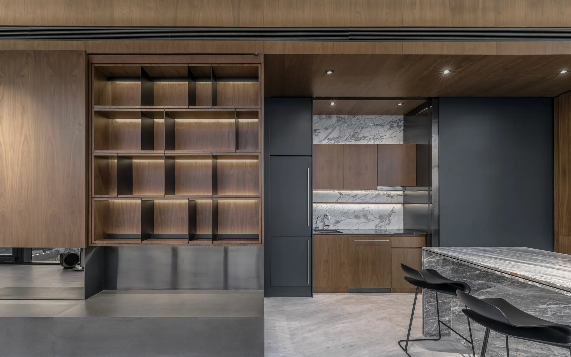 A contemporary kitchen featuring backlit walnut shelves, a marble-clad kitchen island with black bar stools, and dark gray paneled walls.