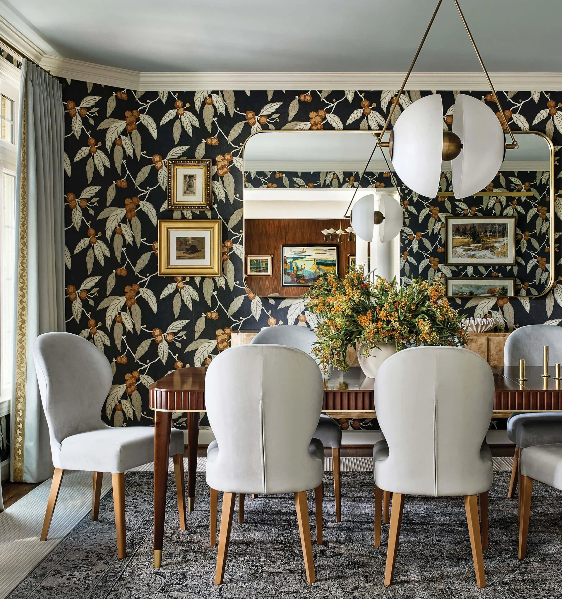 A formal dining room features a dark botanical patterned wallpaper, a wooden dining table surrounded by pale grey upholstered chairs, and a large wall mirror.