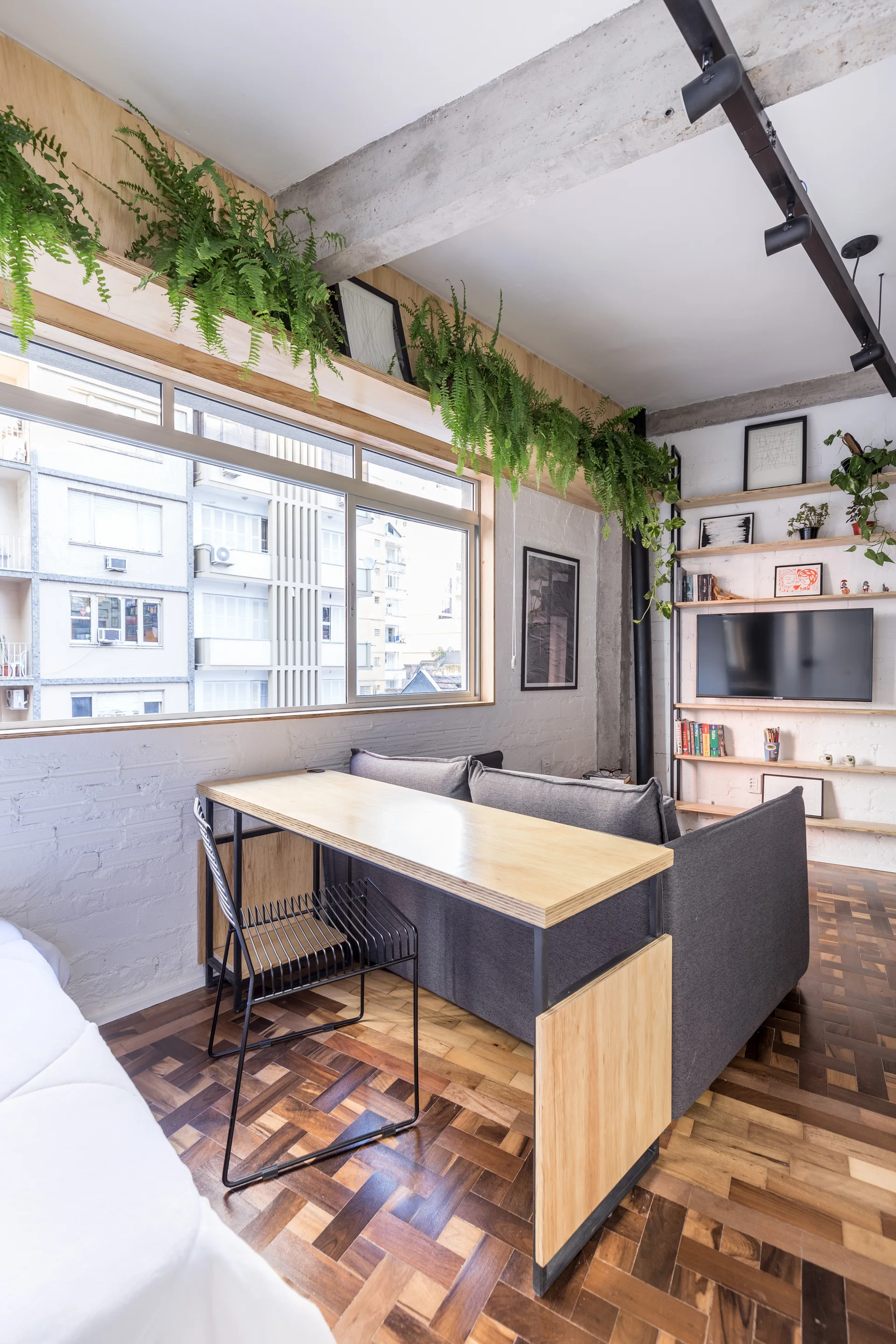 A modern living room featuring a grey sofa with an attached light wood desk, parquet flooring, and ferns hanging above a large window.