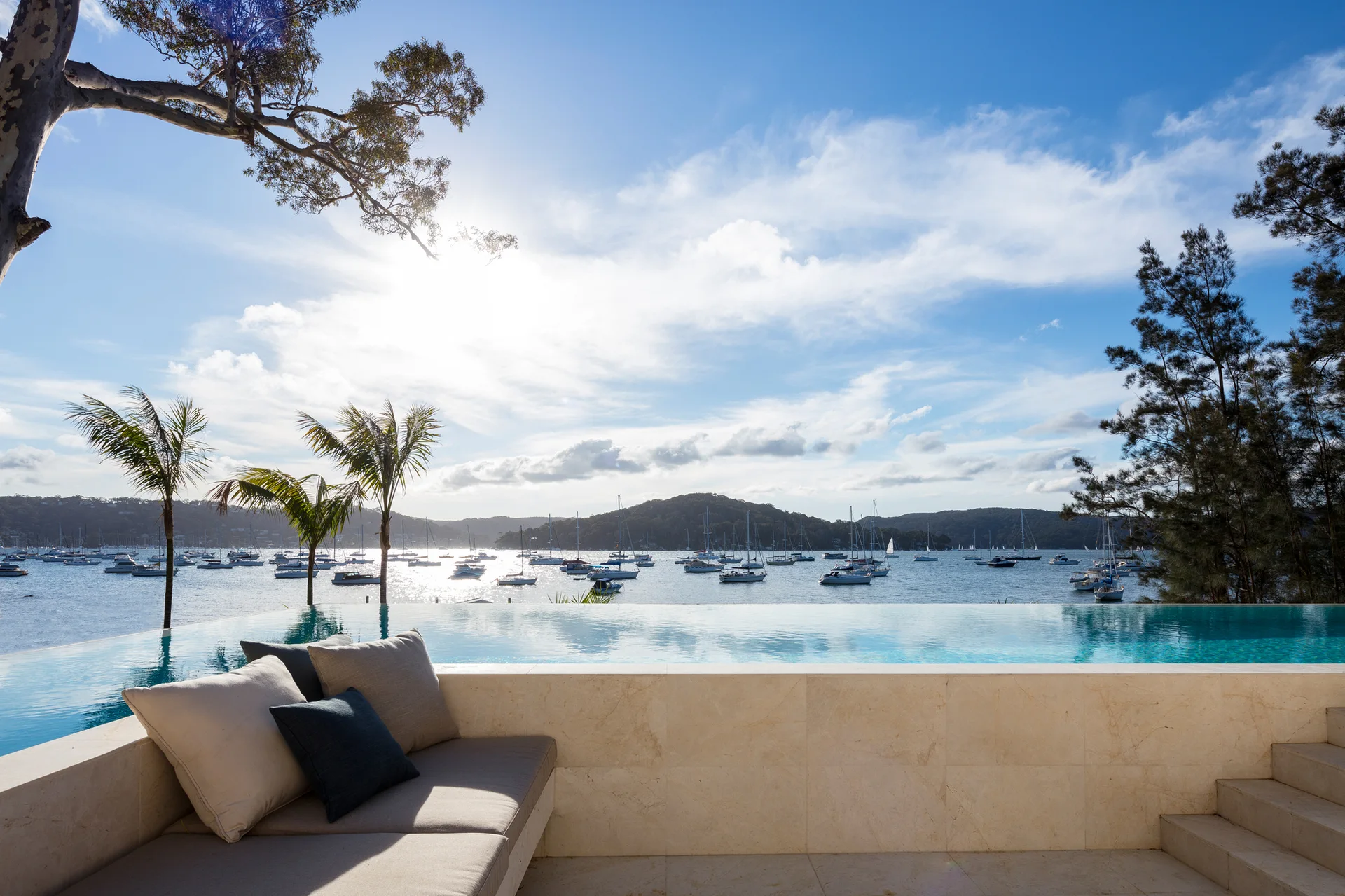 A beige stone built-in outdoor sofa with throw pillows sits next to an infinity pool overlooking a bay filled with sailboats.