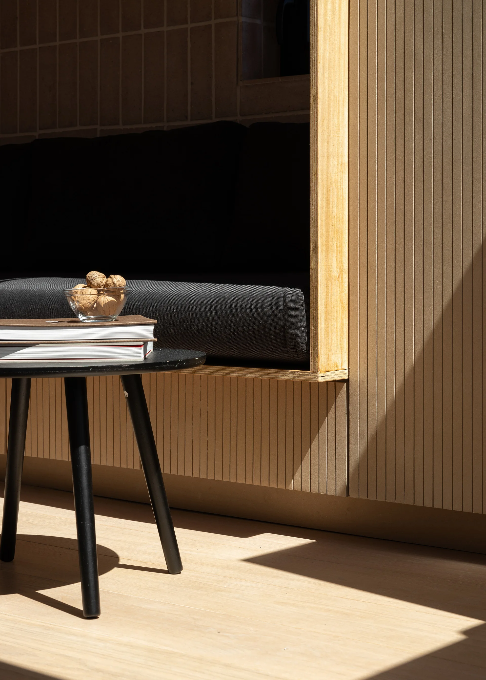 A minimalist built-in wooden bench with charcoal cushions is framed by exposed plywood edges next to a black round side table.