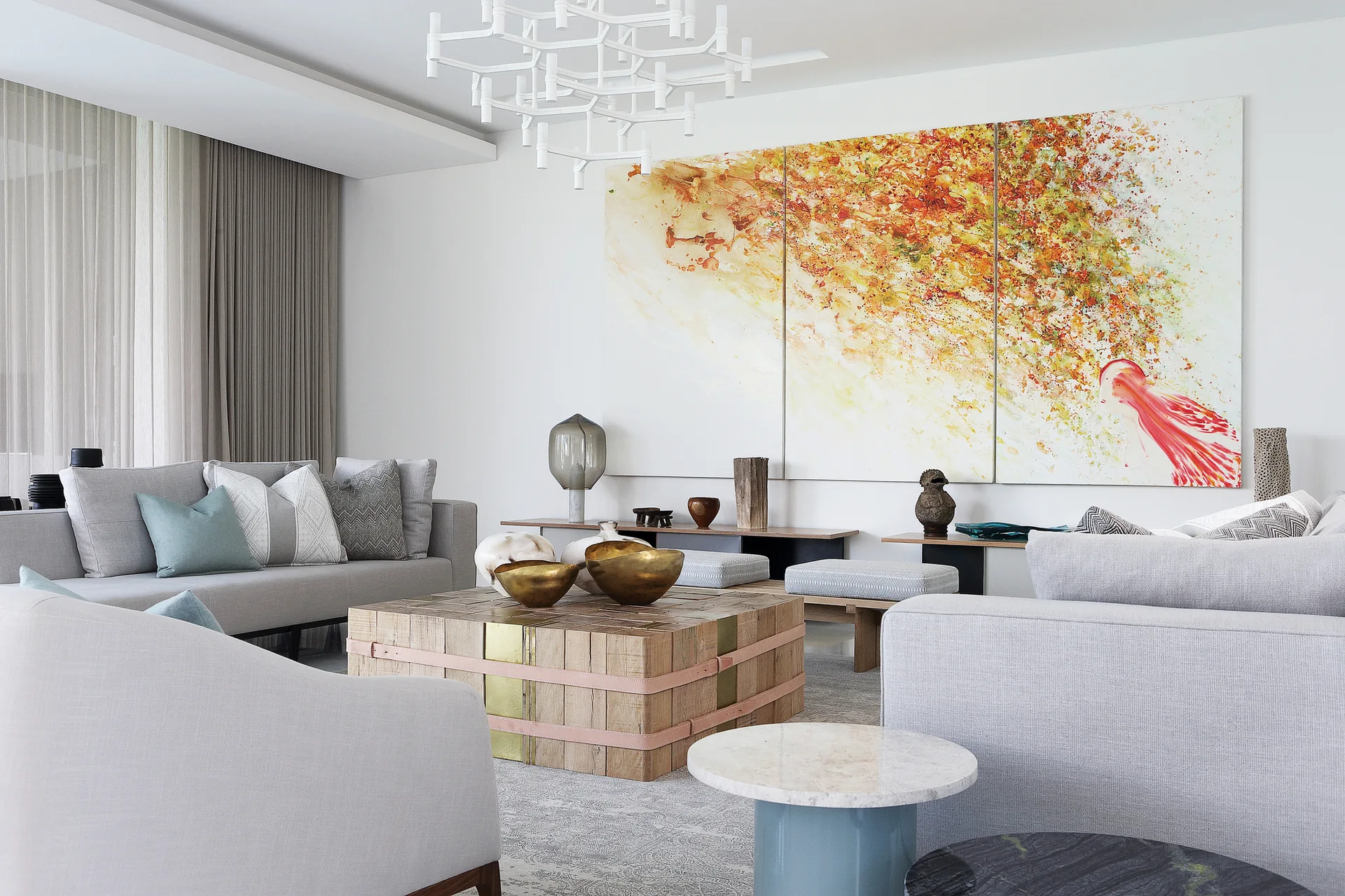A modern living room features light gray sofas, a blocky wood coffee table, and a large three-panel abstract painting on a white wall.