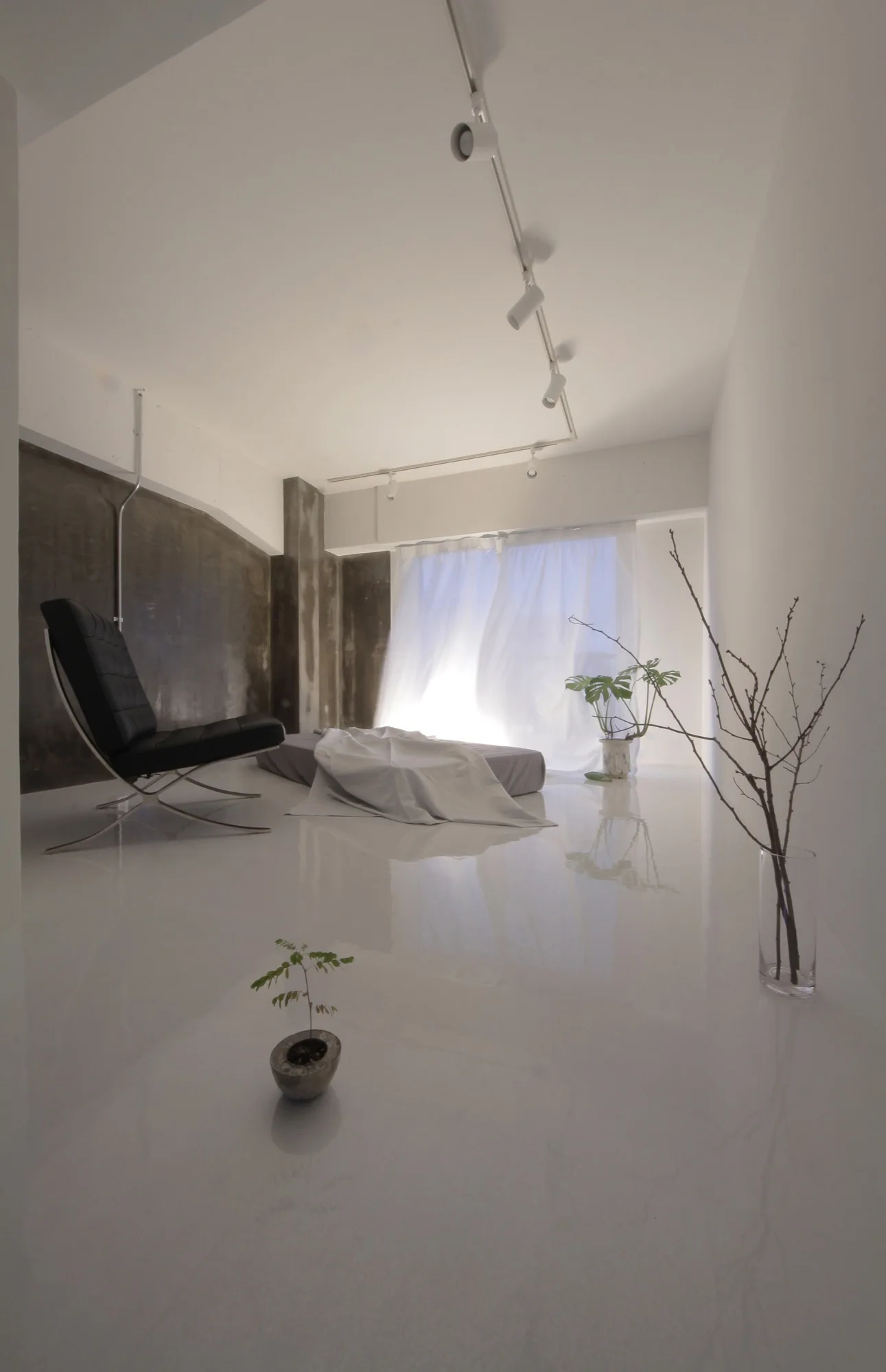 A minimalist room with a highly reflective white floor, a black leather lounge chair, a low daybed, and a raw concrete wall.