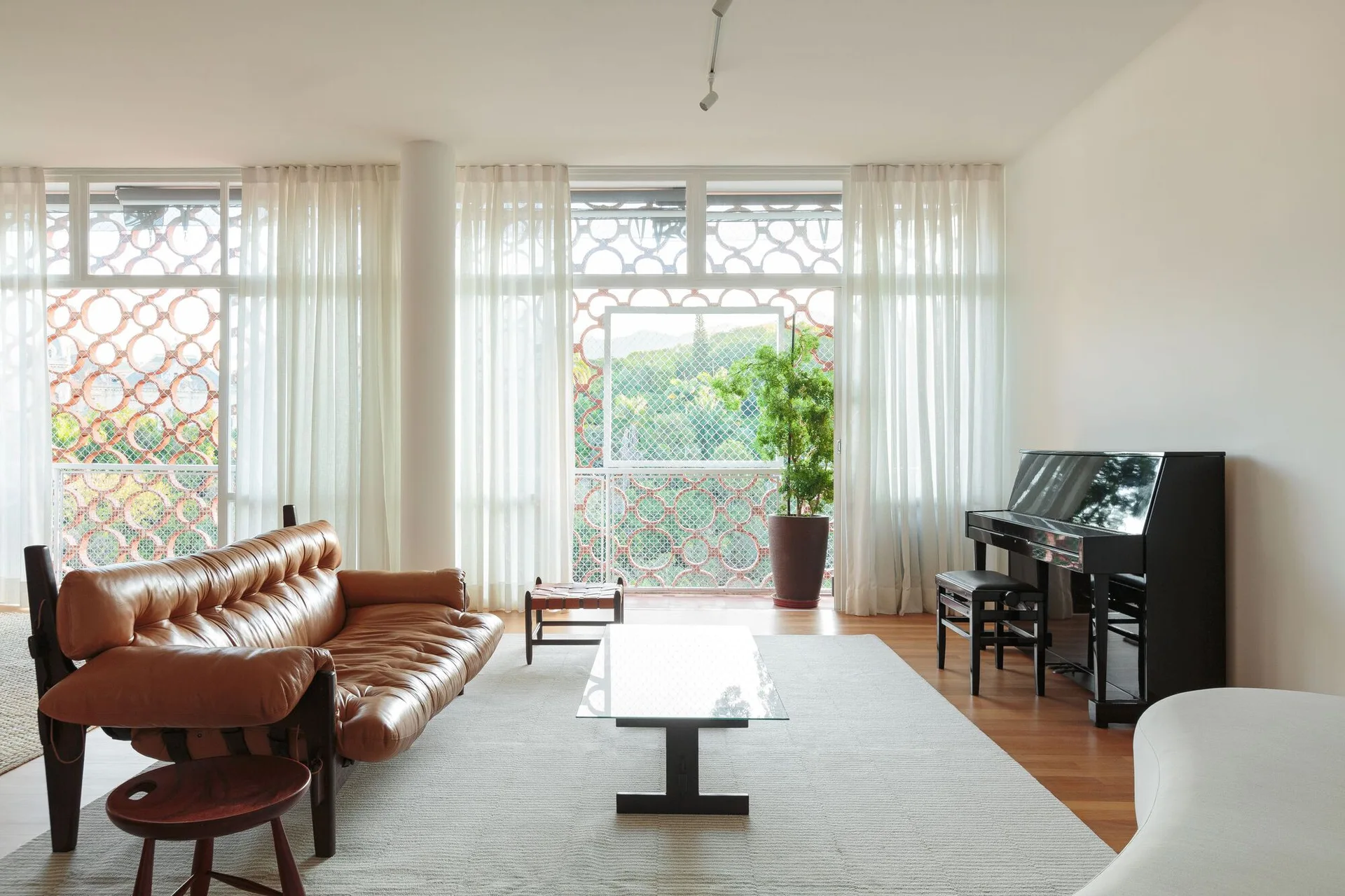 A mid-century living room with a tan tufted leather sofa, glass coffee table, black upright piano, and sheer floor-to-ceiling curtains.