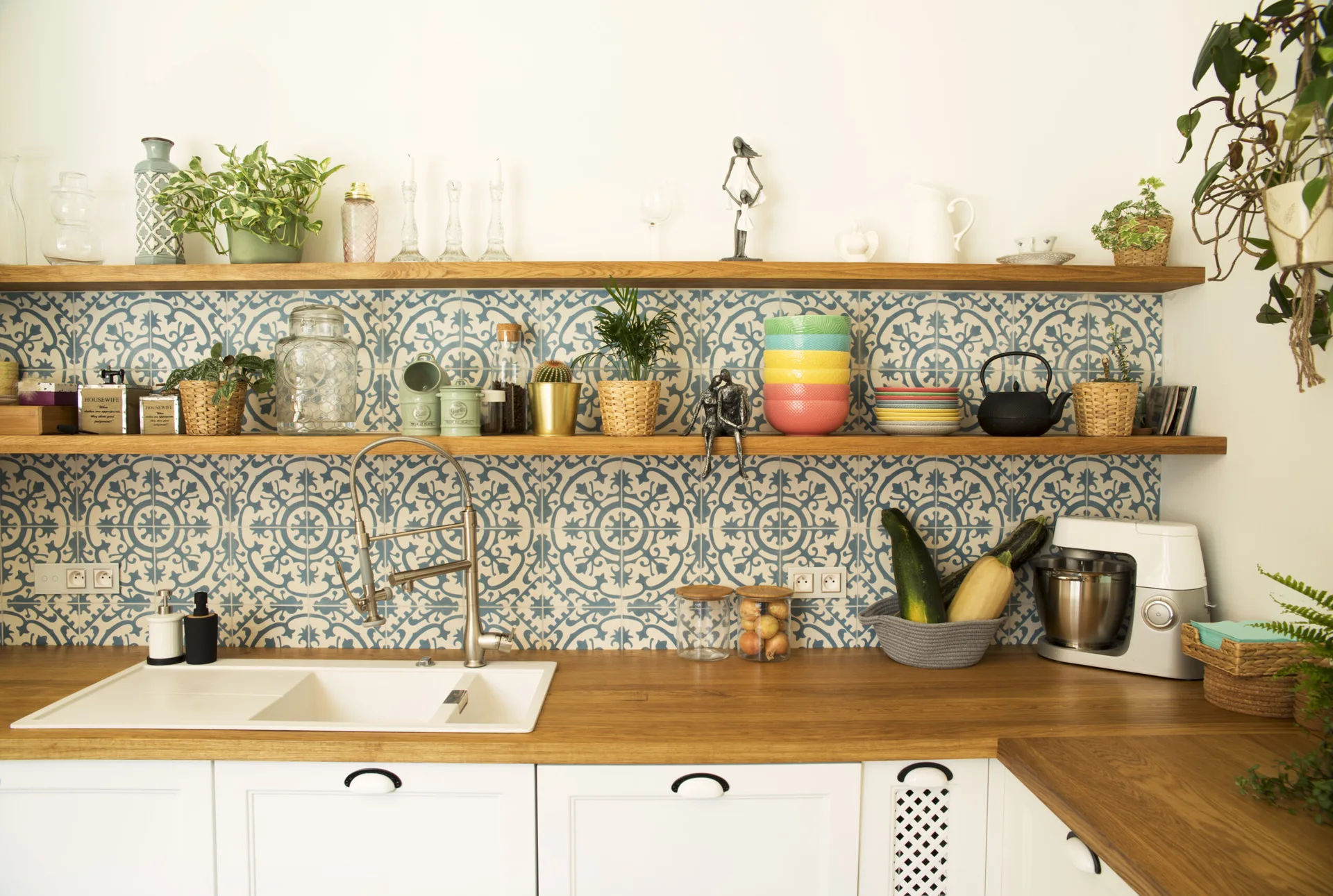 A kitchen featuring a wood countertop, white sink, patterned blue and white tile backsplash, and two wooden open shelves styled with plants and dishes.