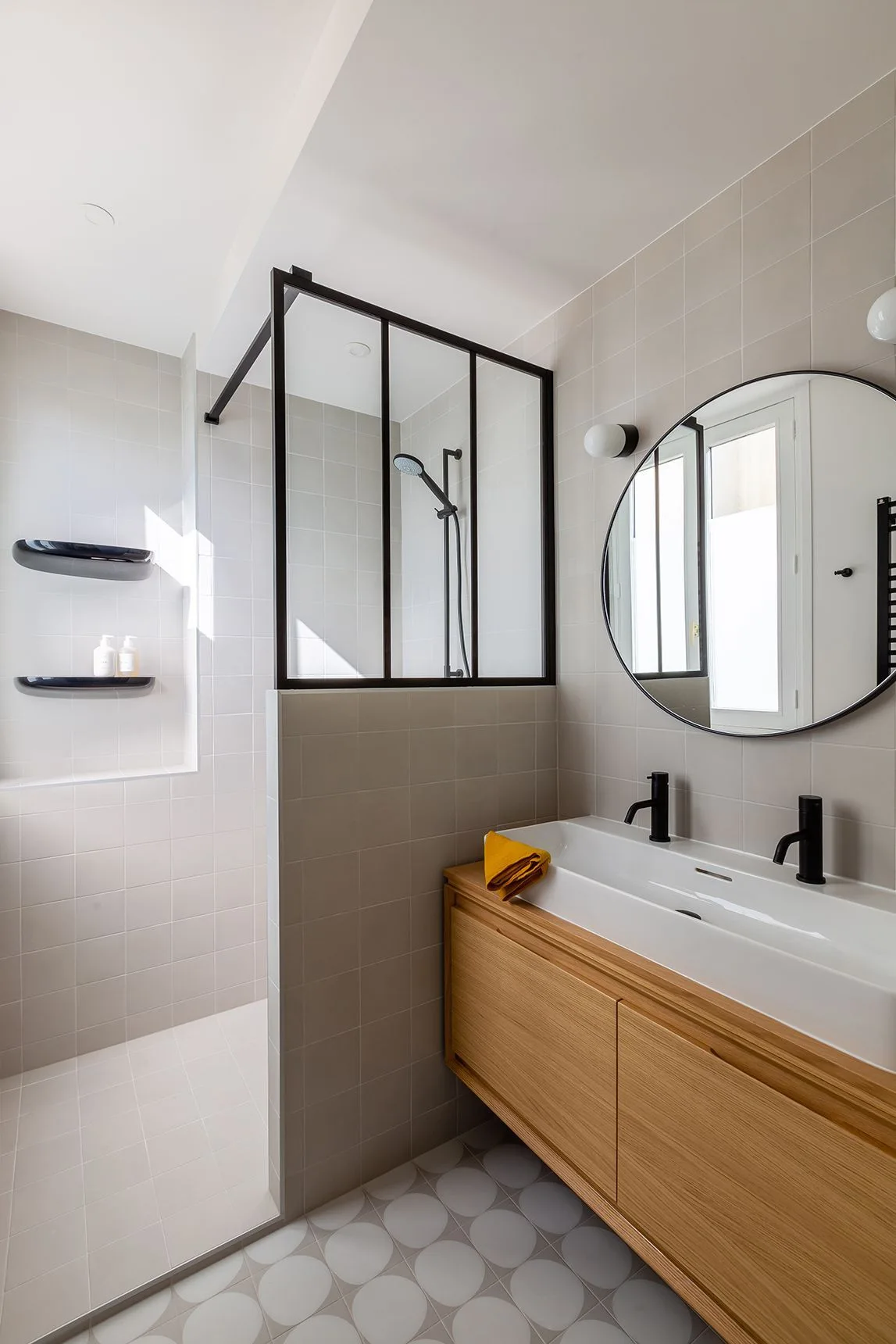 A modern bathroom featuring a floating wood vanity with black faucets, a round mirror, and a walk-in shower with a black-framed glass screen.