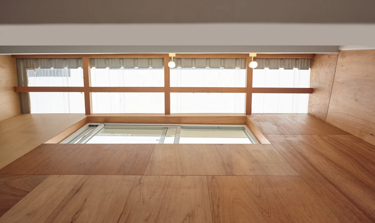 A low-ceilinged loft space with warm plywood paneling, wooden floors, a built-in sliding glass window, and a translucent partition wall.