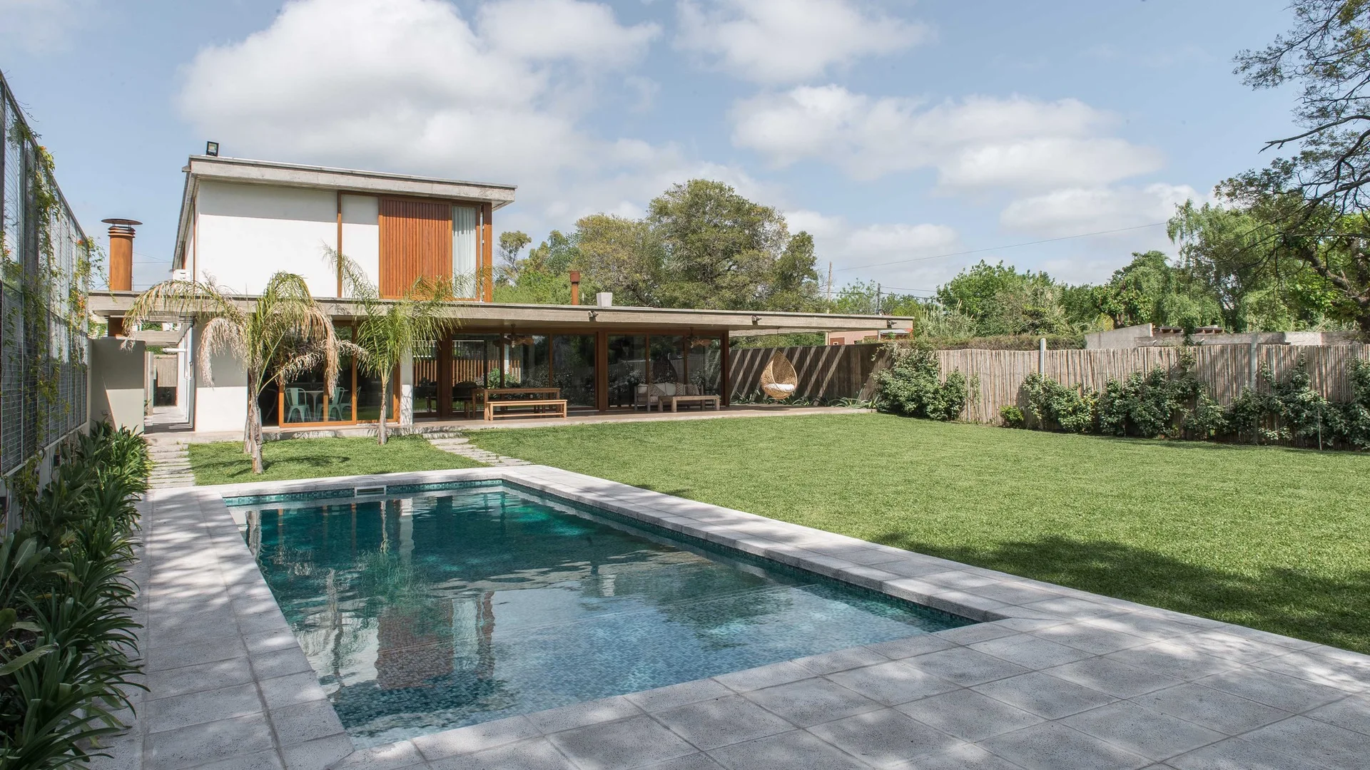 A modern two-story house features a rectangular swimming pool, a large green lawn, a covered concrete terrace, and natural wood facade accents.