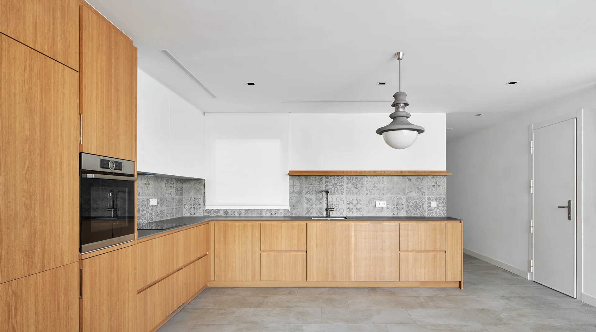 A modern minimalist kitchen features light wood lower cabinets, white upper cabinets, a patterned tile backsplash, and a large grey pendant light.