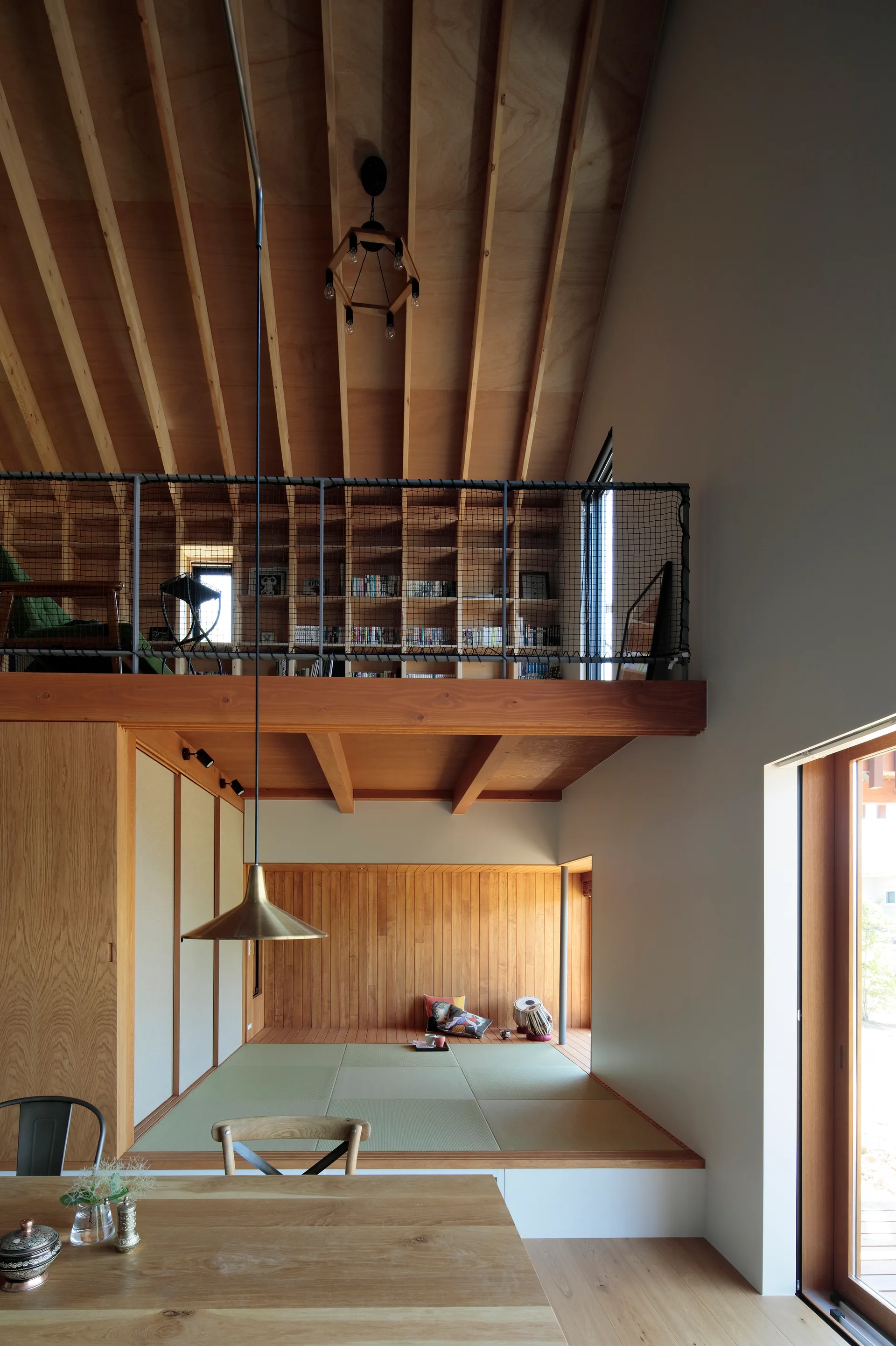 A modern home interior featuring an elevated tatami mat platform below a mezzanine library with built-in wooden bookshelves and safety netting.
