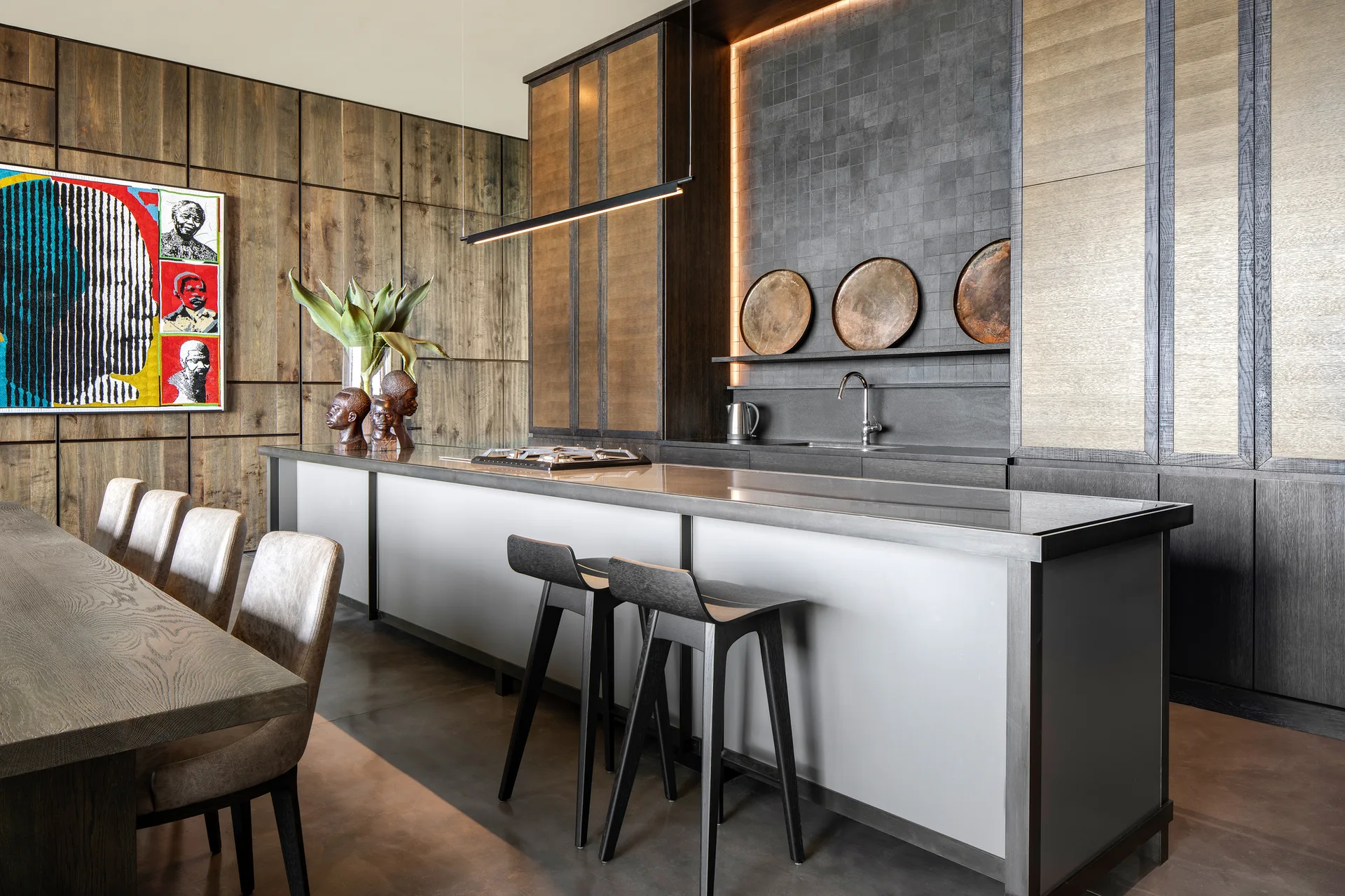 A modern kitchen features dark wood paneled walls, a long island with black bar stools, grey concrete floors, and a dark tiled backsplash.