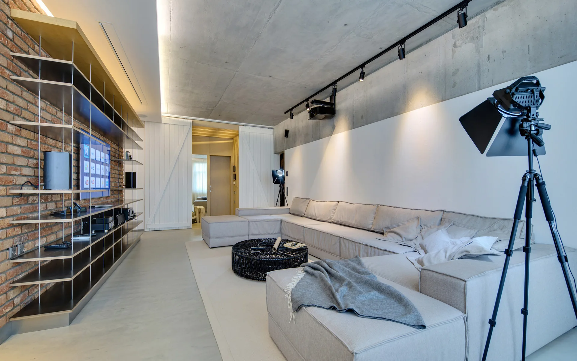 A modern living room featuring a large grey sectional sofa, an exposed concrete ceiling with track lighting, and a red brick wall with built-in shelves.