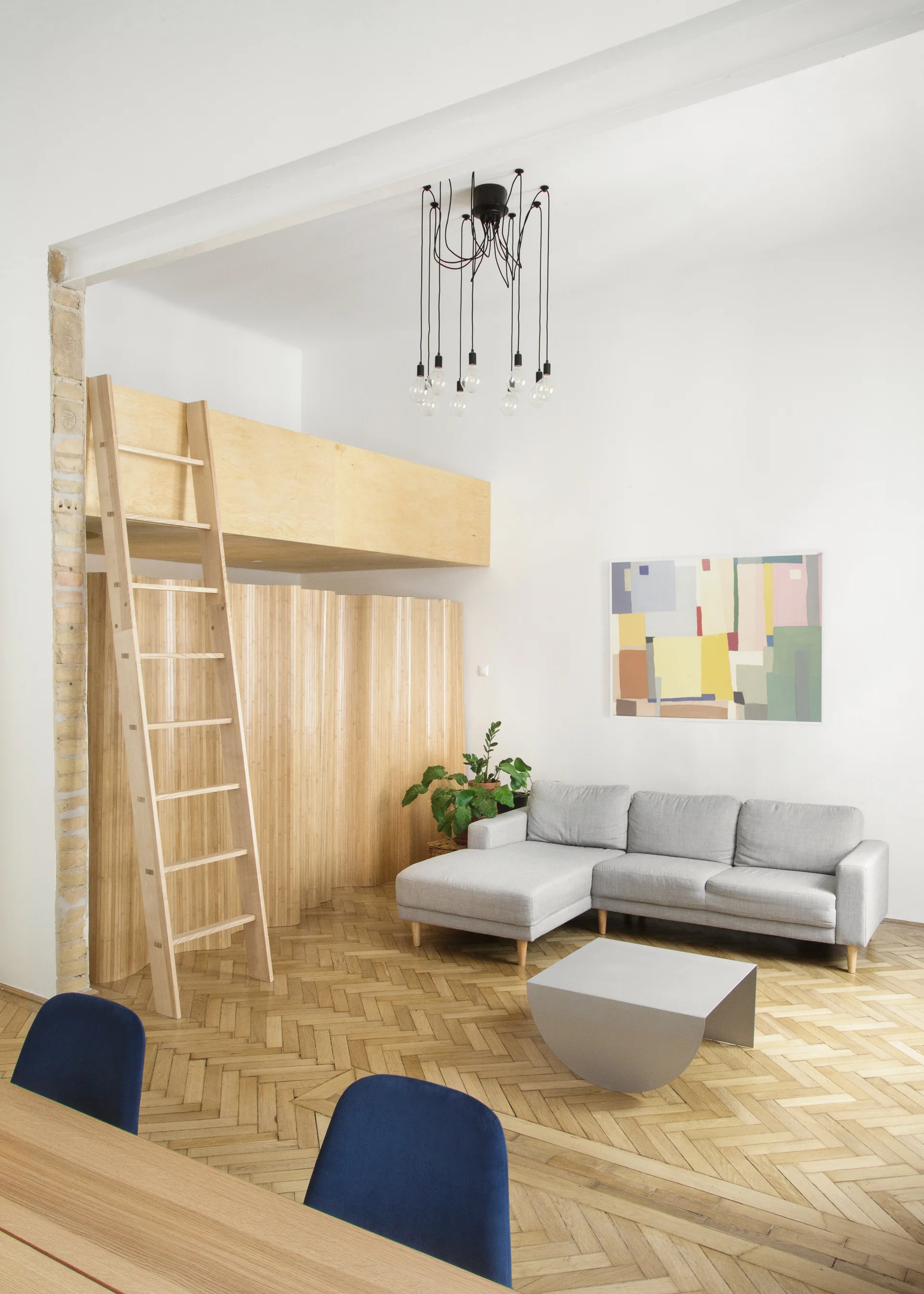 A light wood mezzanine loft with a ladder overlooks a grey sectional sofa, sculptural coffee table, and herringbone wood floor in a white living room.