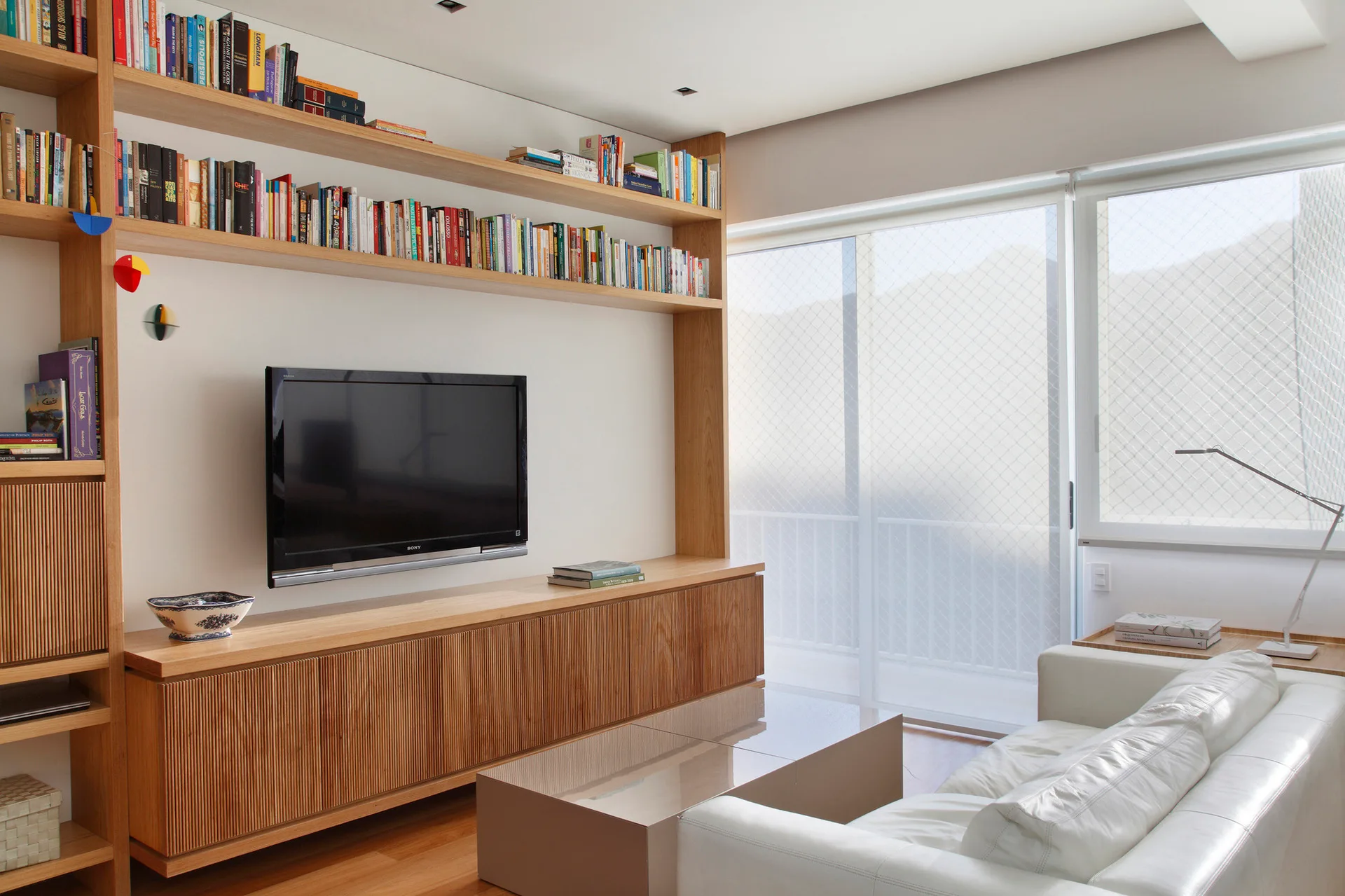 A modern living room featuring a custom wood wall unit with fluted lower cabinets and open bookshelves, paired with a white leather sofa.