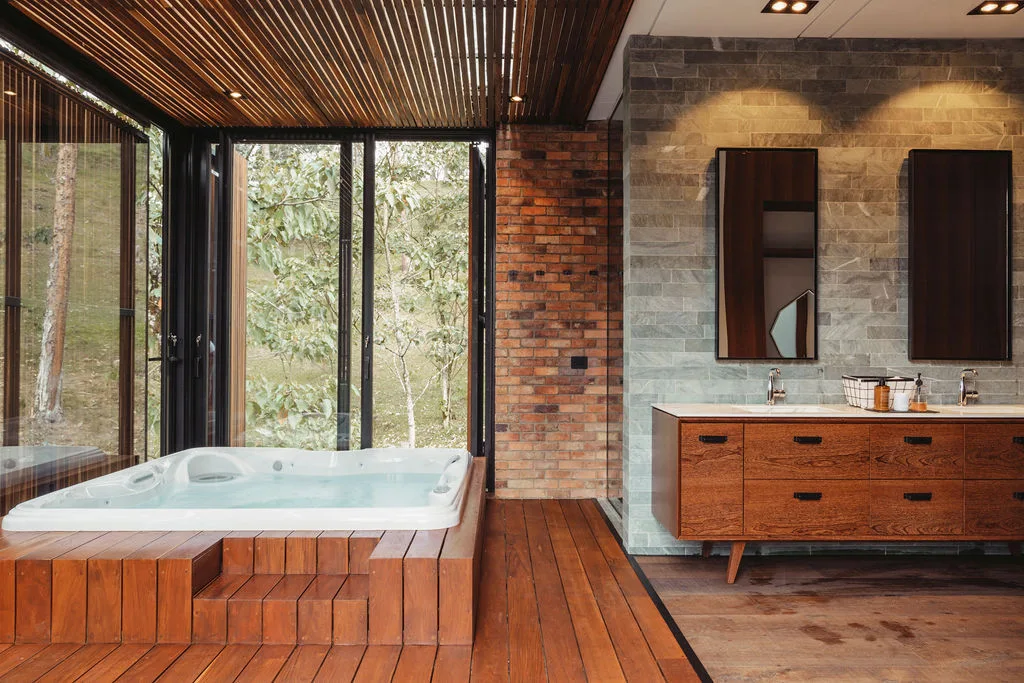 A spa bathroom features a wooden-enclosed jacuzzi tub next to floor-to-ceiling windows, alongside a double vanity set against a grey stone tiled wall.