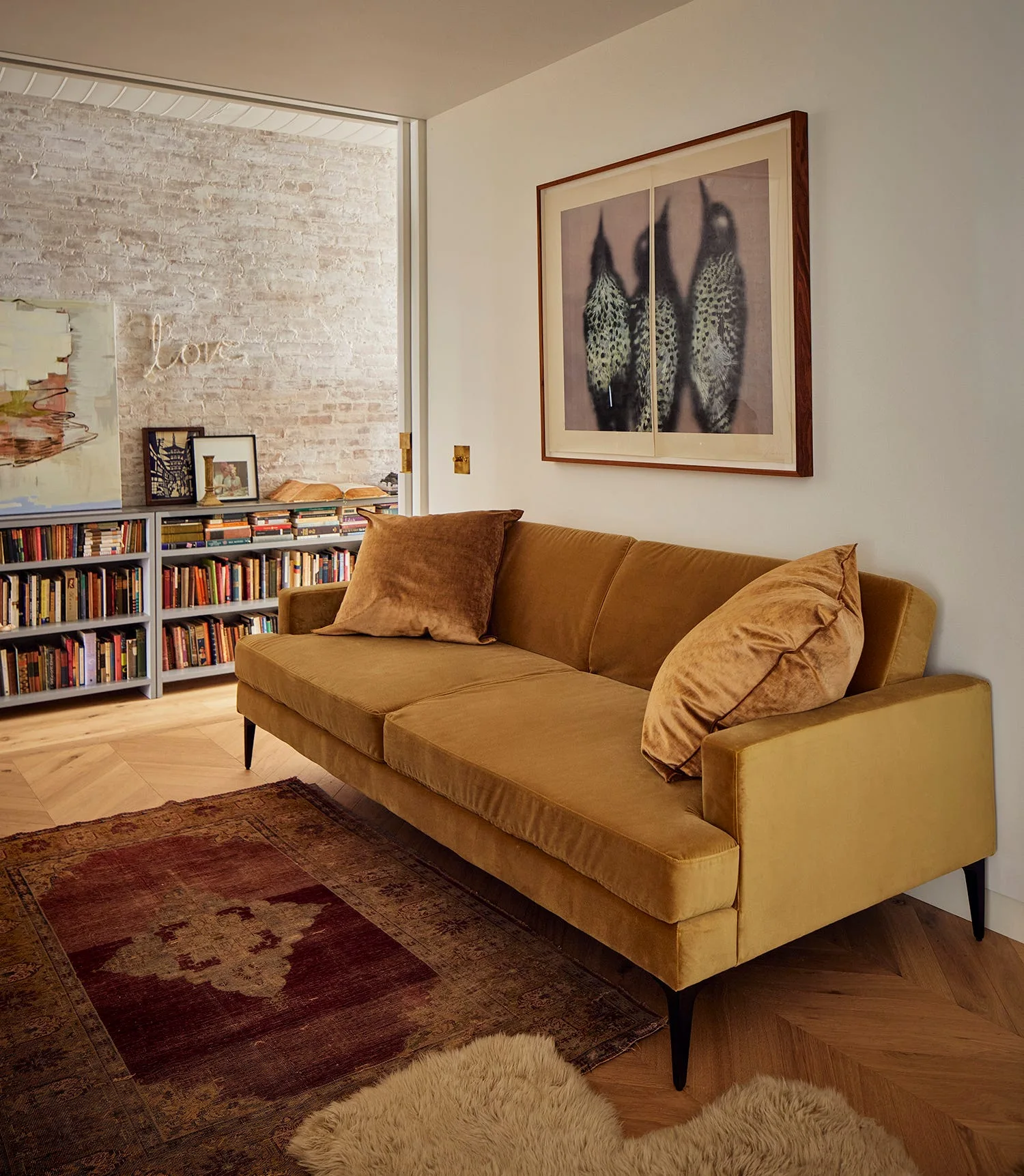 A mustard yellow velvet sofa sits on a herringbone wood floor in front of an off-white wall decorated with a large framed bird print.