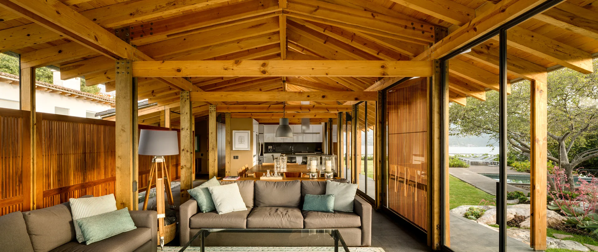 A modern living room featuring a vaulted wood-beam ceiling, grey sofas, and floor-to-ceiling glass walls that open to an outdoor garden.
