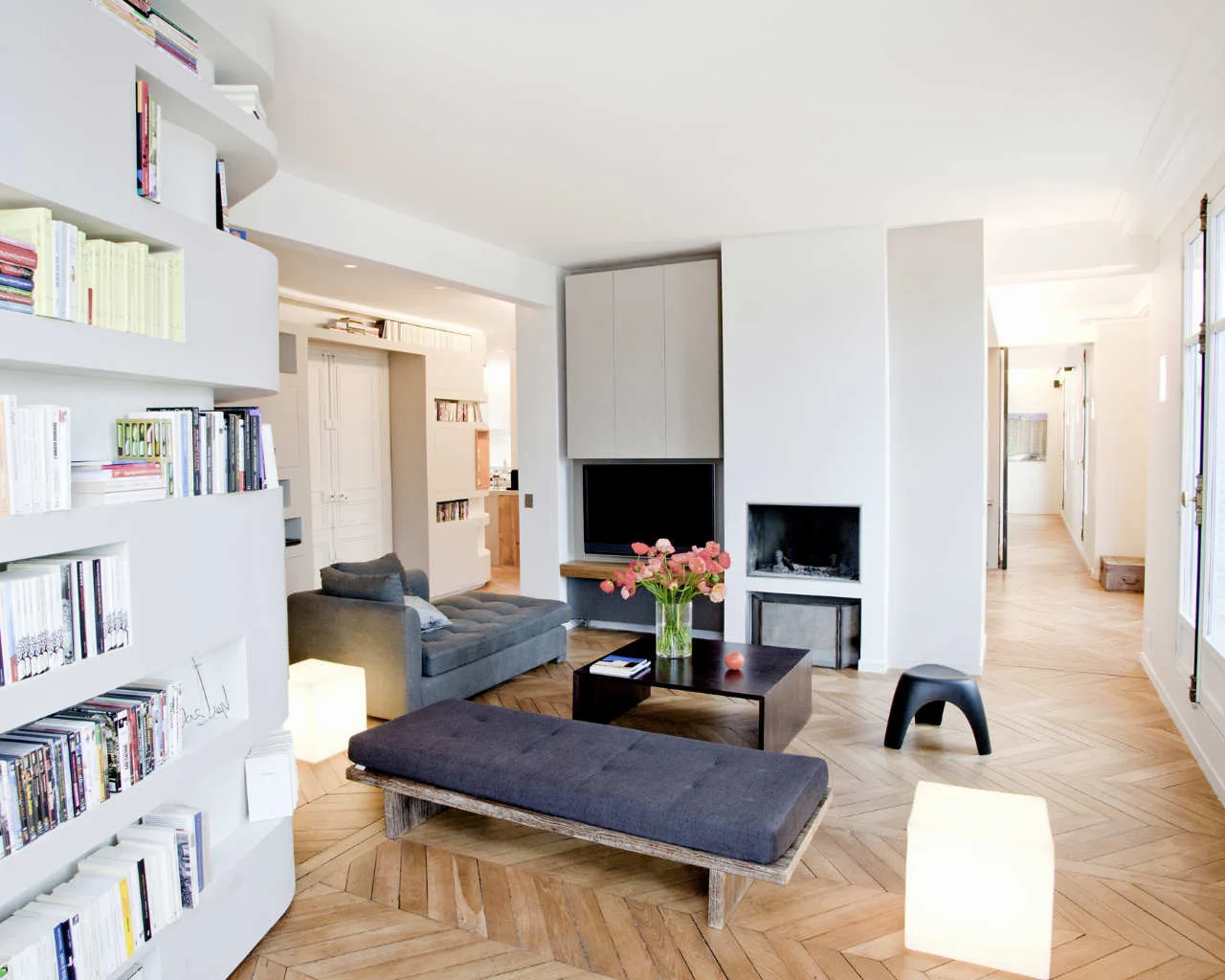 A contemporary living room featuring white curved built-in shelves, a gray sofa, a low bench, and light wood herringbone parquet flooring.