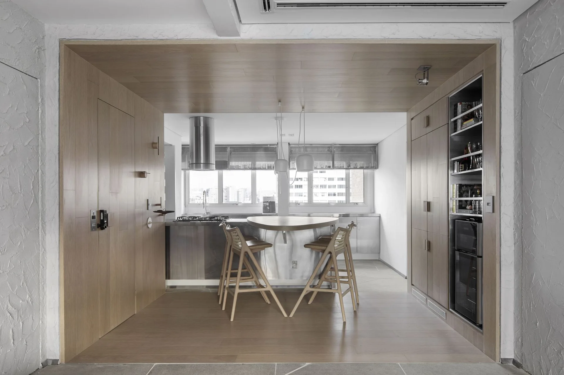 A modern kitchen set inside a wood-paneled alcove features a stainless steel island with wooden stools, integrated cabinetry, and hanging pendant lights.