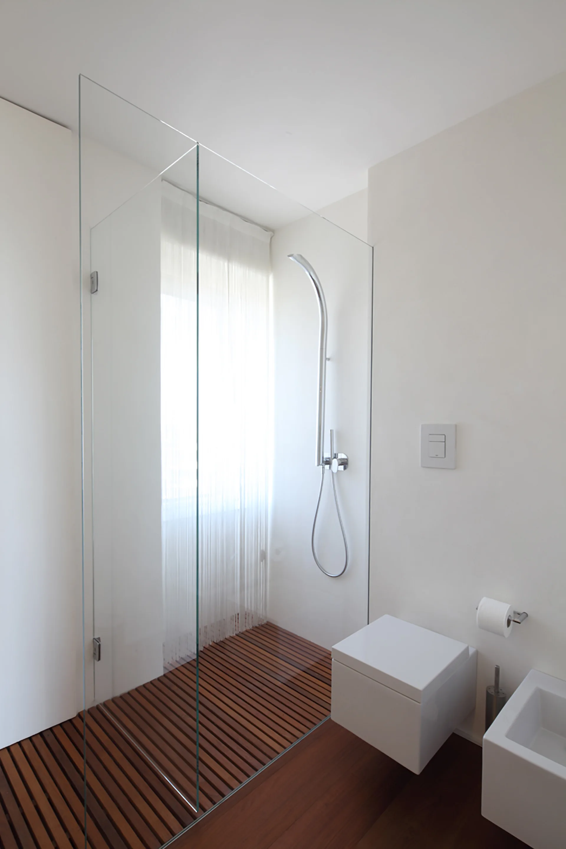 A minimalist bathroom features a glass walk-in shower with wooden slats on the floor alongside a wall-mounted white toilet.