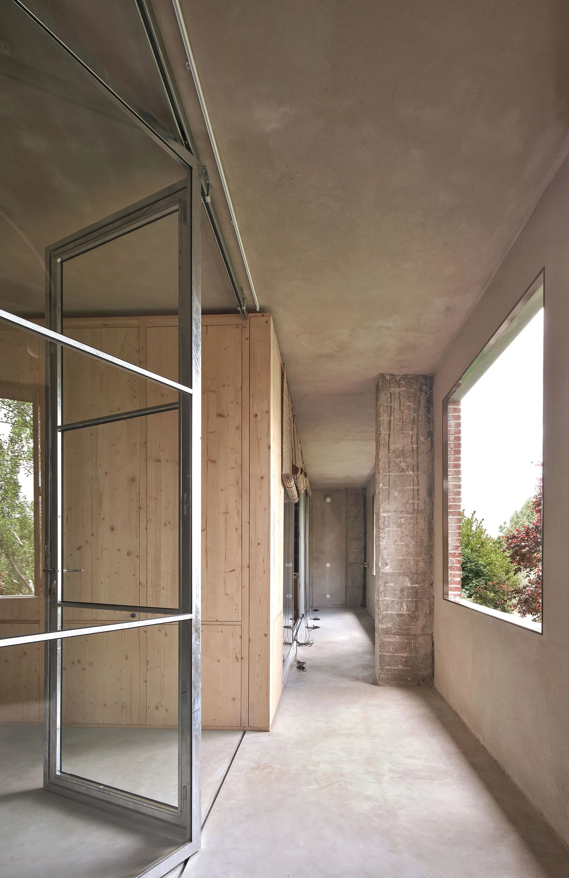 A minimalist concrete hallway features a tall light wood cabinet wall, a rough concrete pillar, and a large metal-framed glass sliding door.