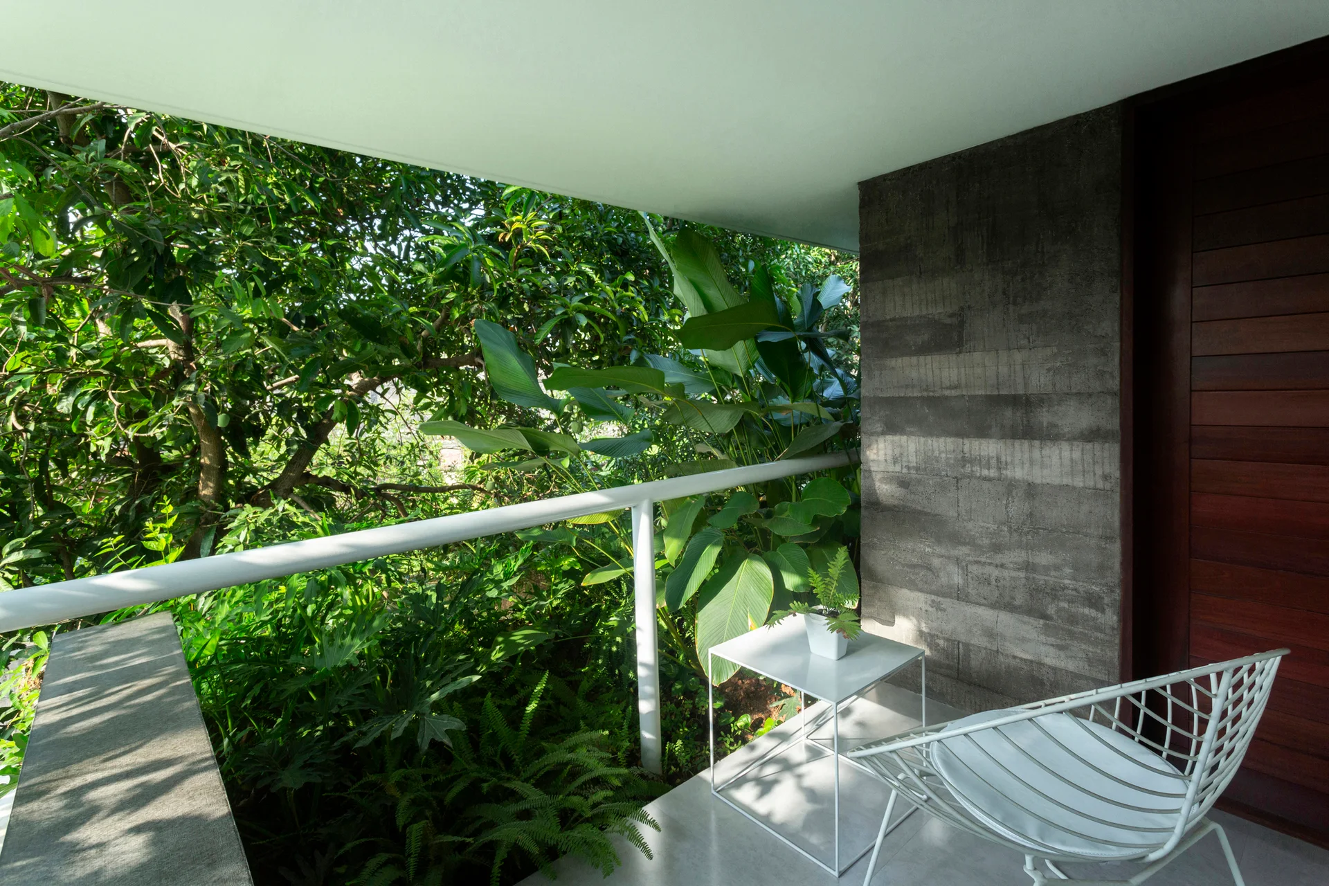 A modern balcony features a white wire metal chair and side table set against an exposed concrete wall and lush green garden view.