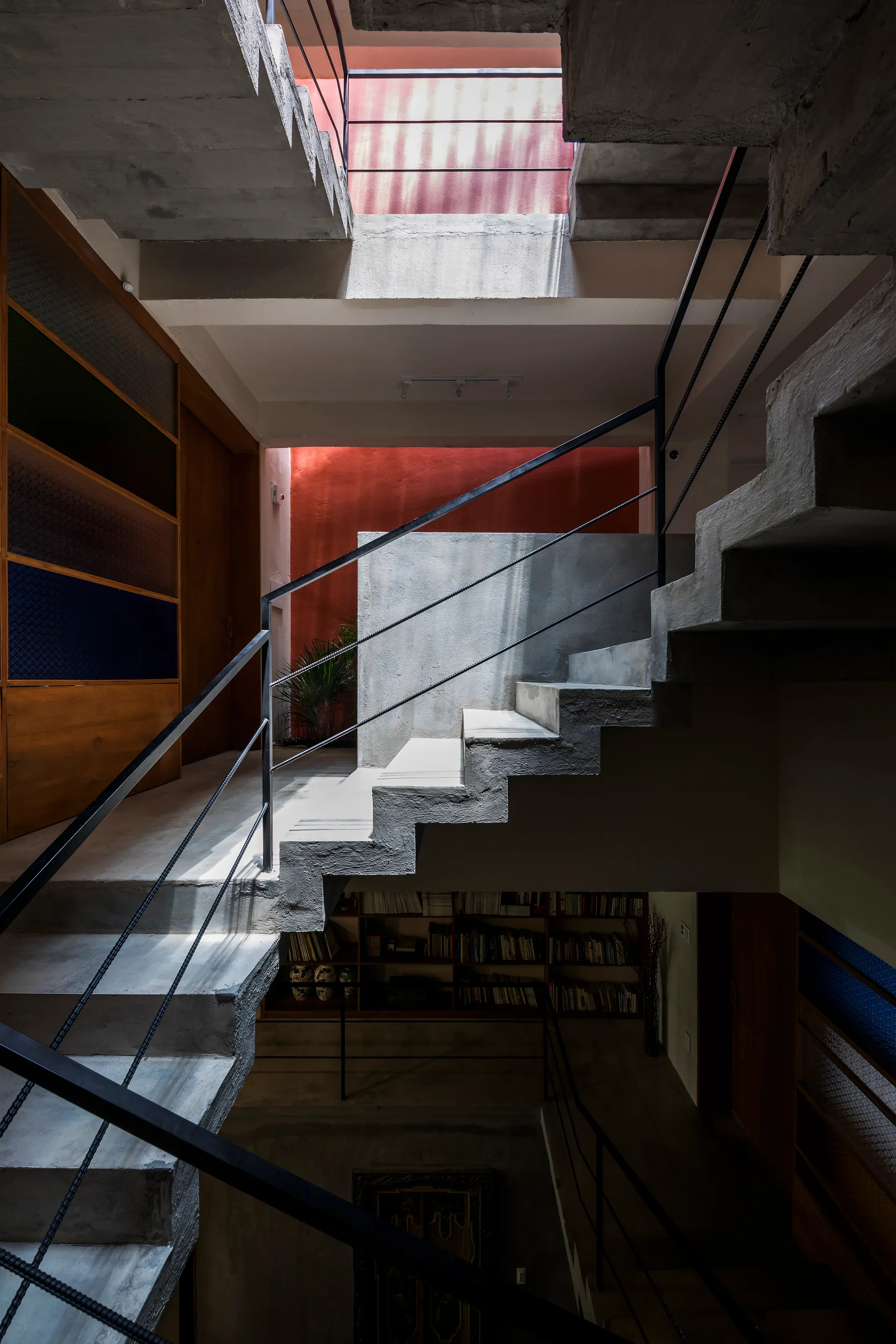A multi-level interior featuring a raw concrete staircase with black metal railings next to a vibrant orange accent wall illuminated by natural light.