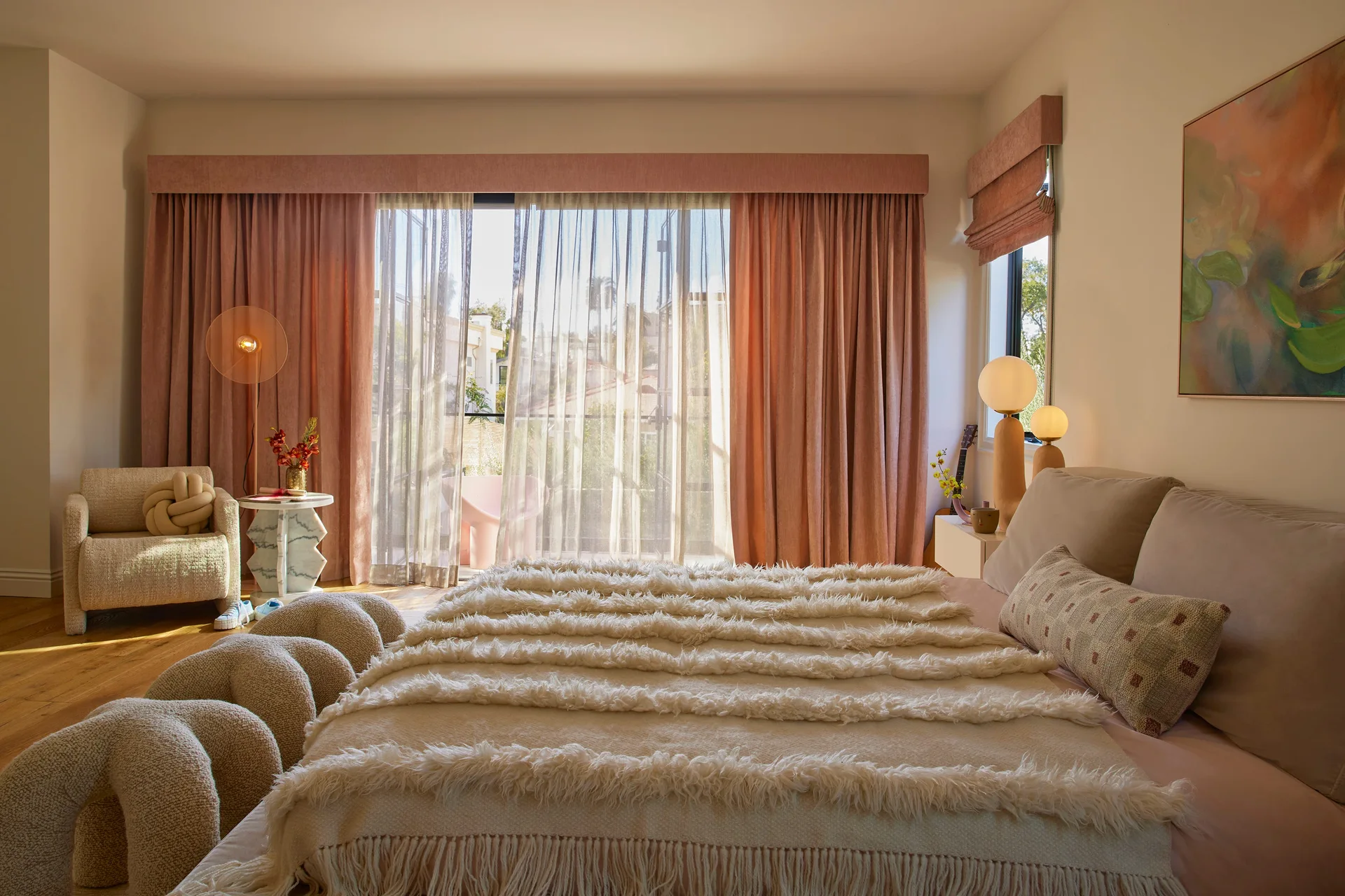 A contemporary bedroom features a bed with a fringed shag throw, long pink drapes over sliding glass doors, and a sculptural armchair.