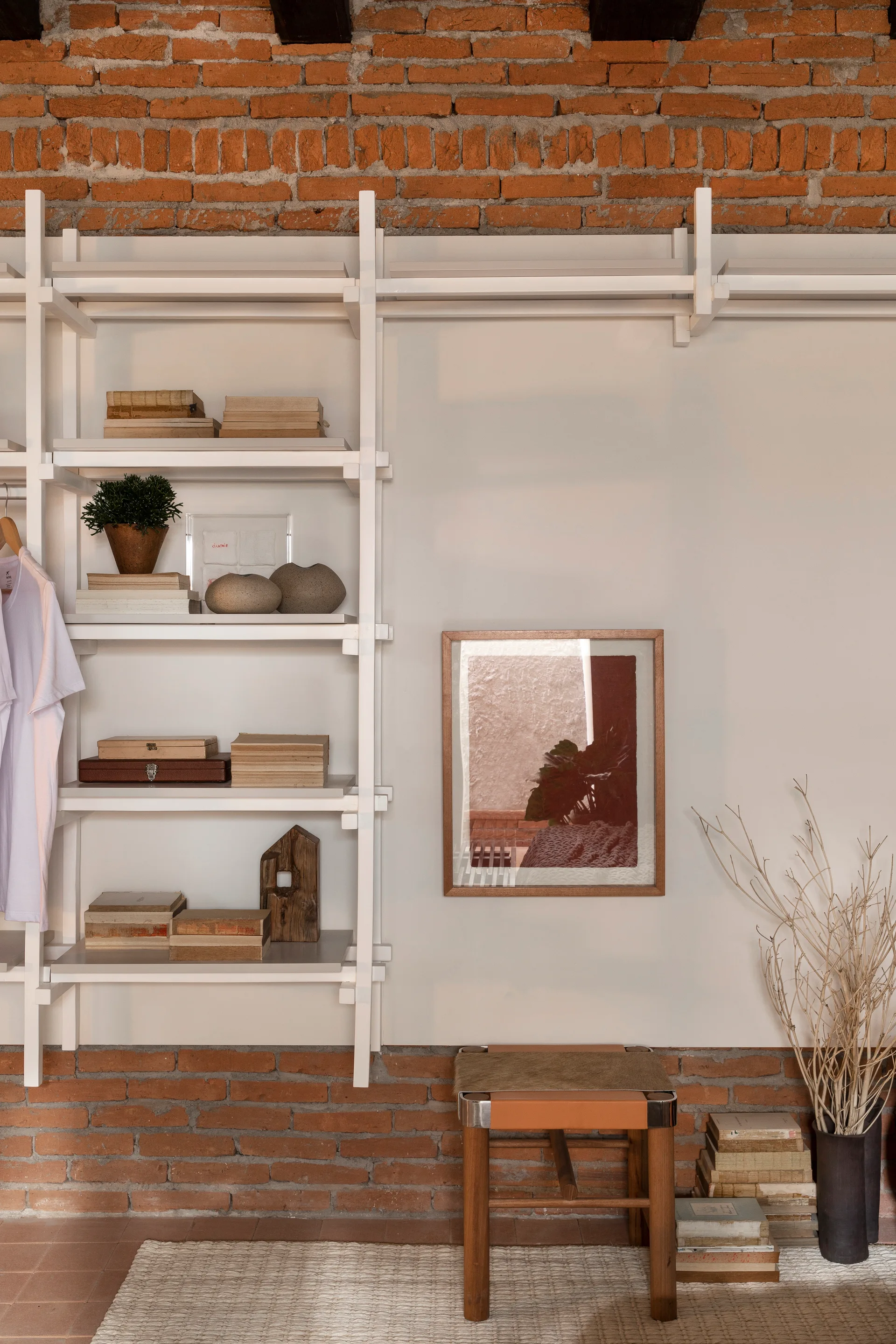 A modular white shelving unit with books and decor is mounted on a wall with exposed red brick next to a framed picture and a wooden stool.