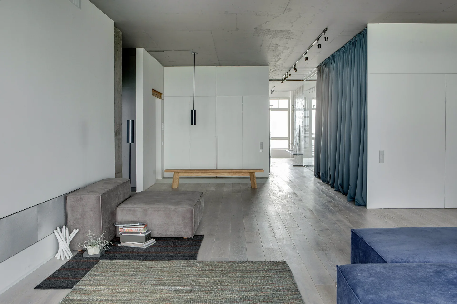 A minimalist living room features a raw concrete ceiling, light wood flooring, a gray modular sofa, and a floor-to-ceiling blue-gray curtain.
