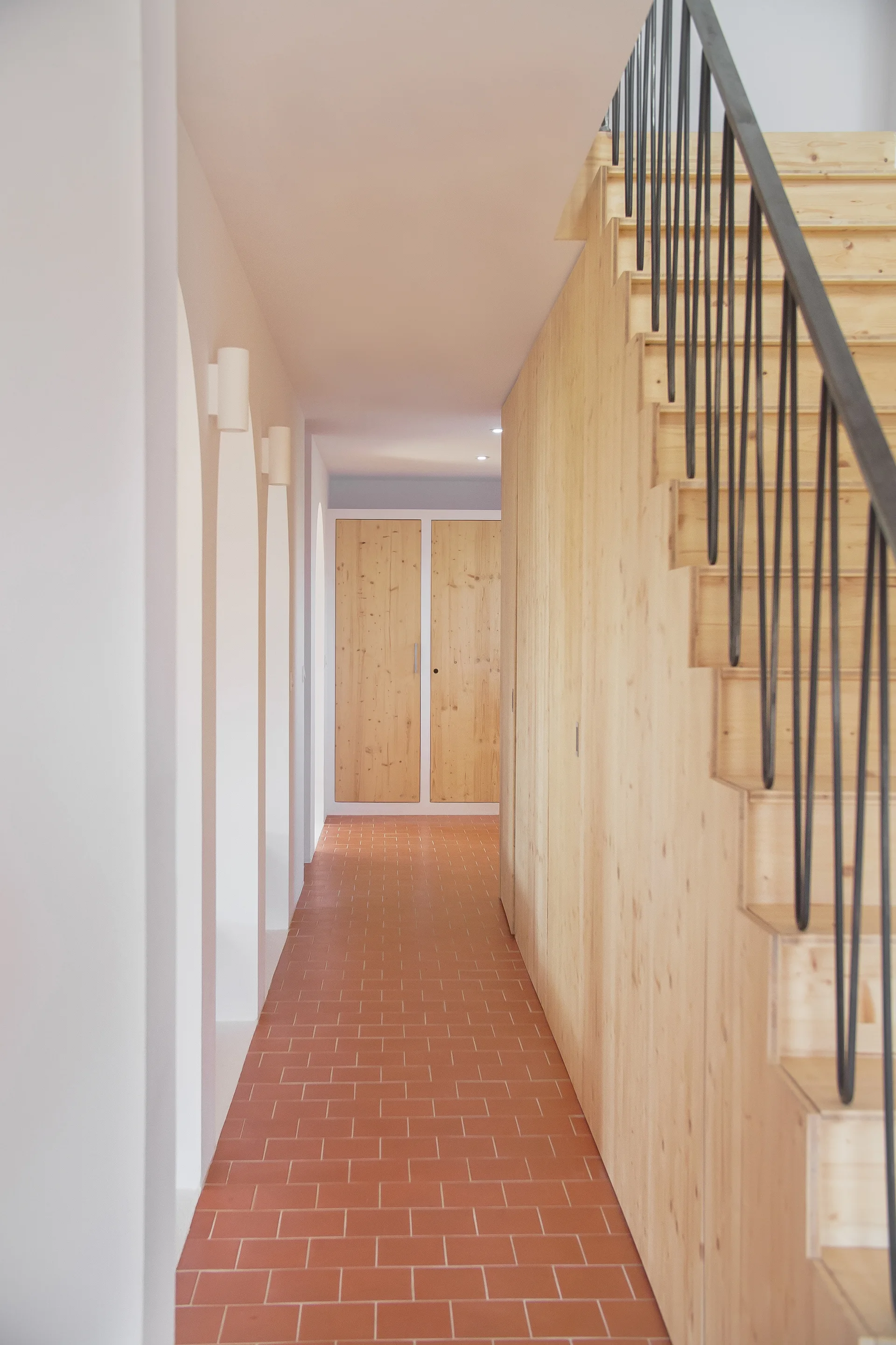 A minimalist hallway features a terracotta tile floor, a light wood paneled staircase with black metal railing, and white arched wall alcoves.