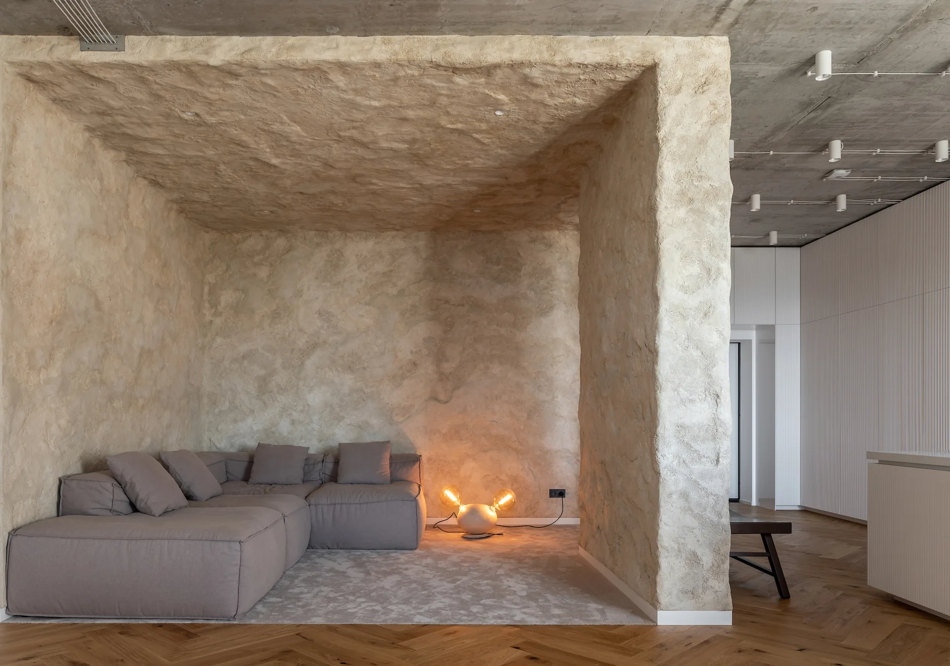 A modular gray sectional sofa sits inside a textured beige plaster alcove with a low-glowing floor lamp next to herringbone wood floors.