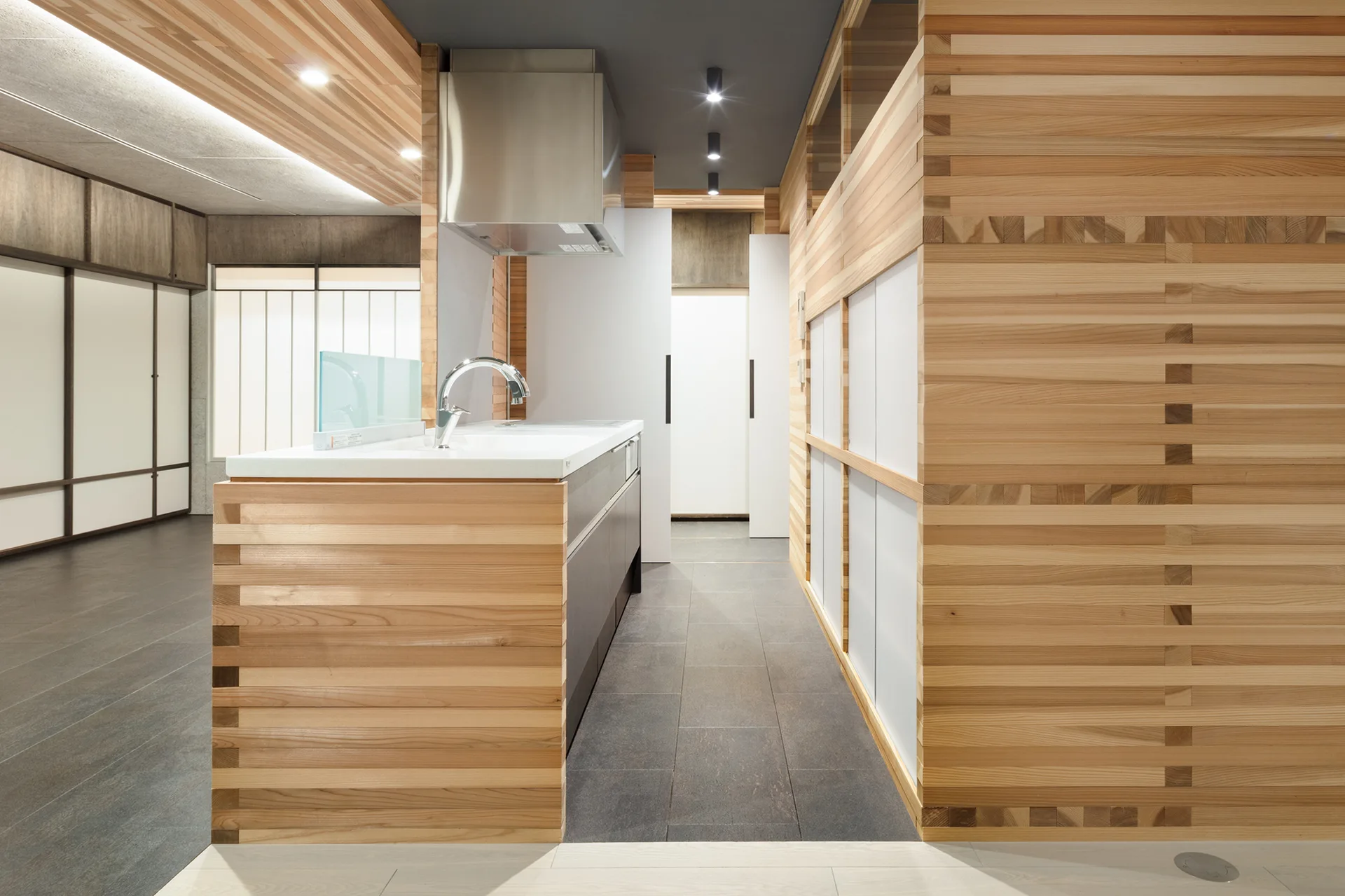 A minimalist kitchen features a central island and walls clad in horizontal light wood slats, dark gray tile flooring, and a stainless steel faucet.