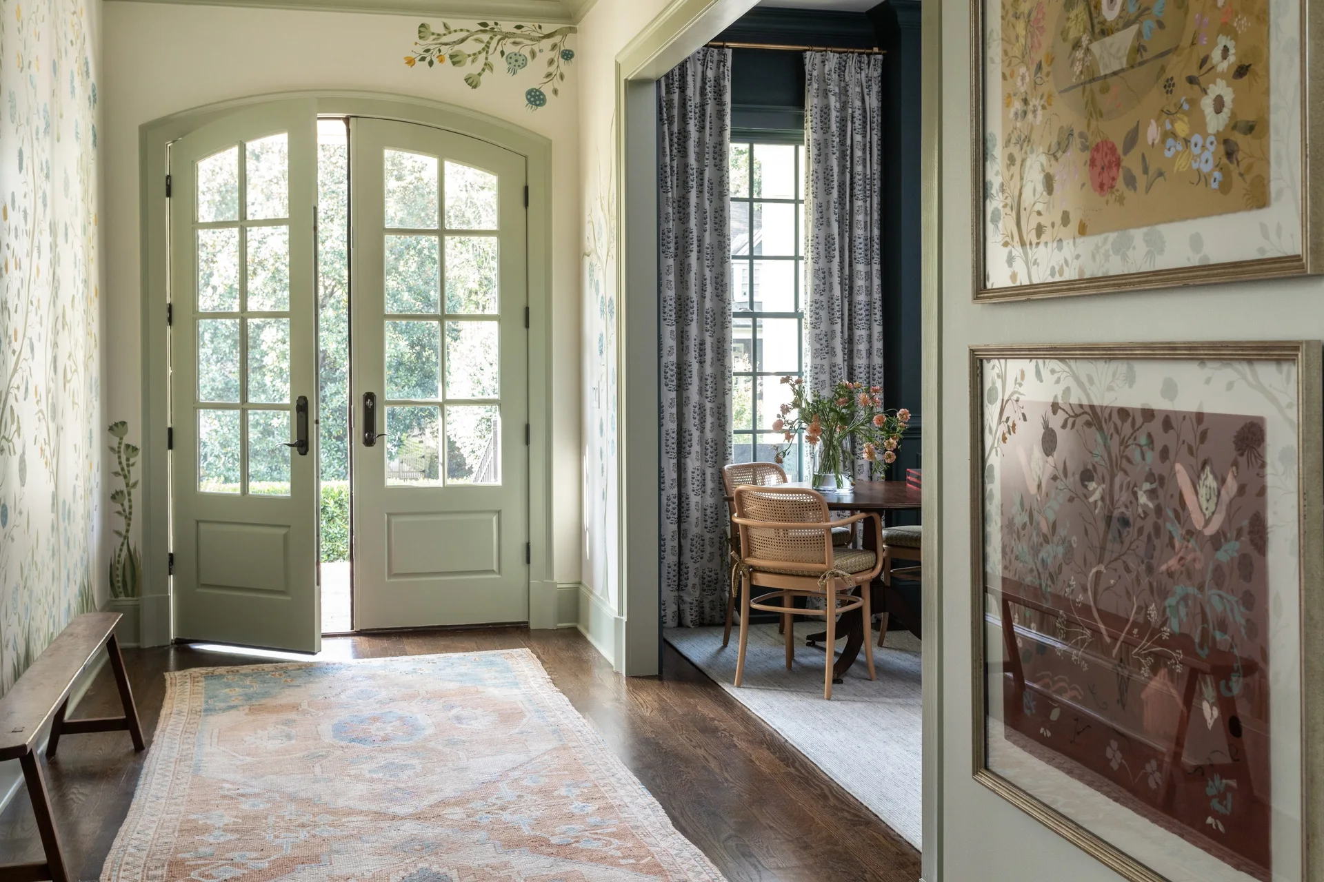 A bright foyer features light green arched double doors, floral wallpaper, an ornate rug, and an opening into a dining room with cane chairs.
