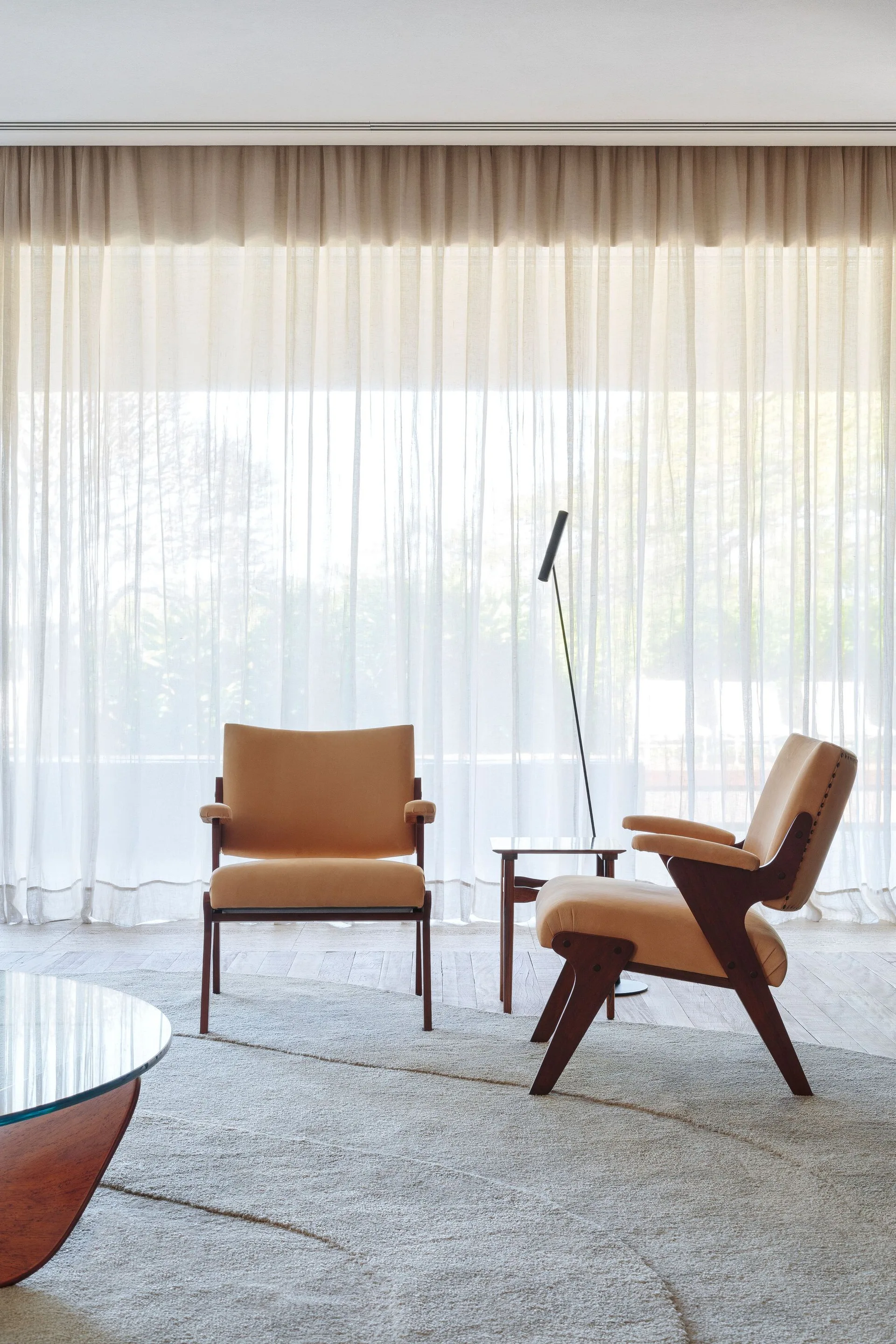 Two tan mid-century modern armchairs sit on a textured beige rug in front of a floor-to-ceiling sheer curtain with a small side table and slim floor lamp.