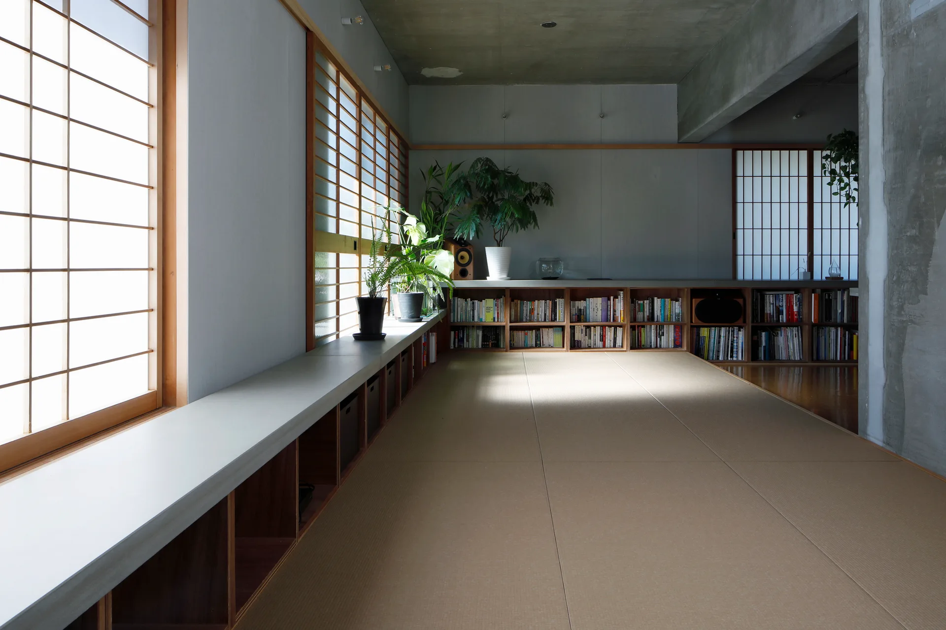 A minimalist room features tatami mat flooring, a long built-in wooden bookshelf, and shoji screen sliding windows.