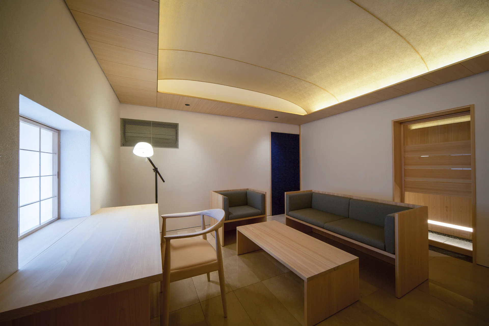A minimalist lounge features a curved backlit ceiling, a light wood built-in desk by a grid window, a wooden sofa with gray cushions, and tile flooring.
