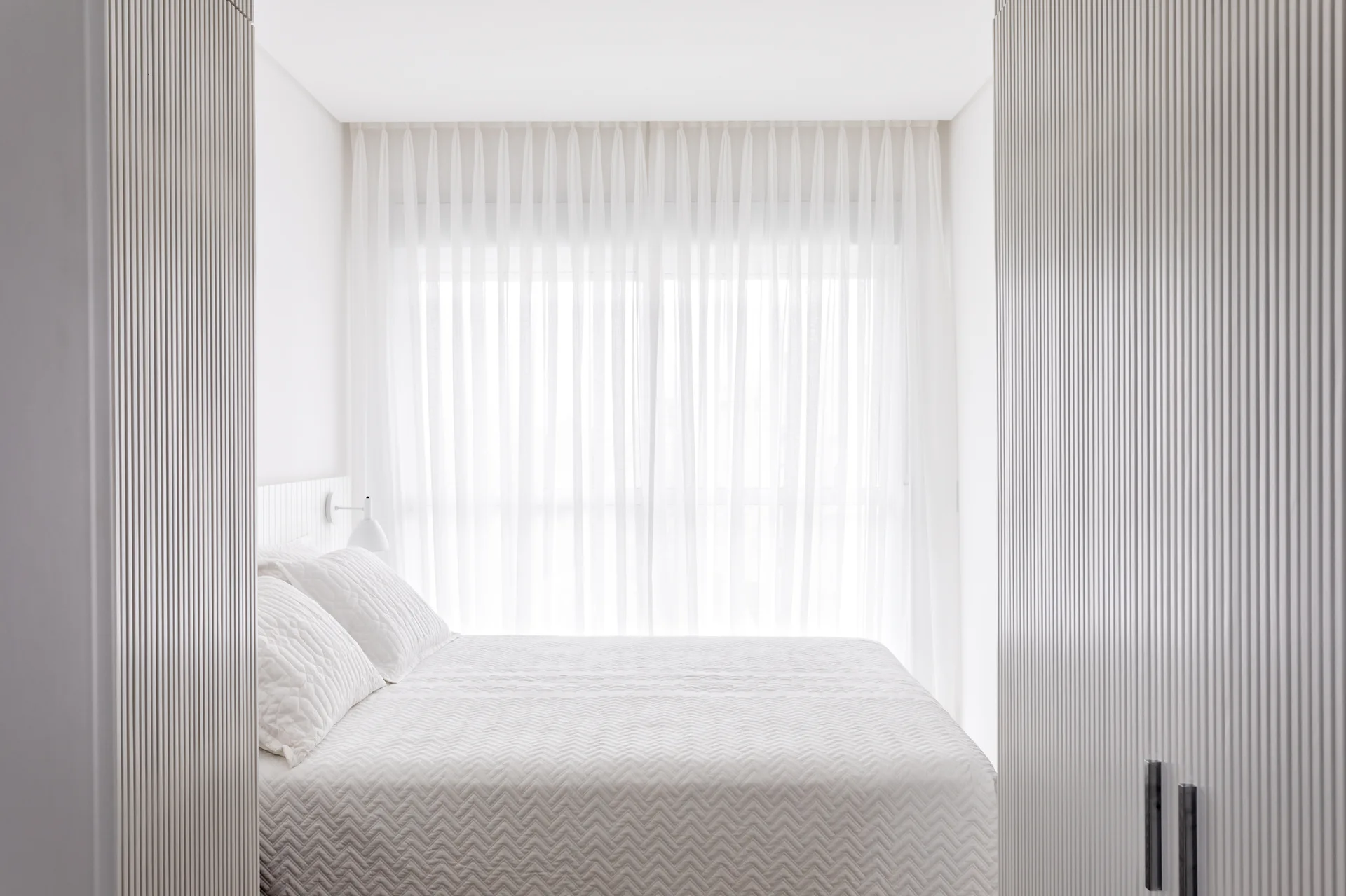 A minimalist white bedroom featuring a bed with quilted bedding, framed by vertical fluted wall panels and soft sheer curtains.