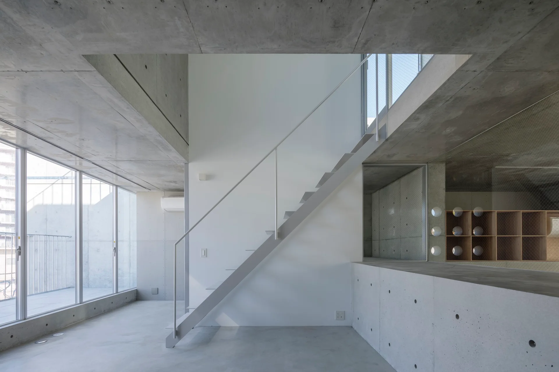 A minimalist room with exposed concrete ceilings and floors features a white metal staircase alongside large windows.
