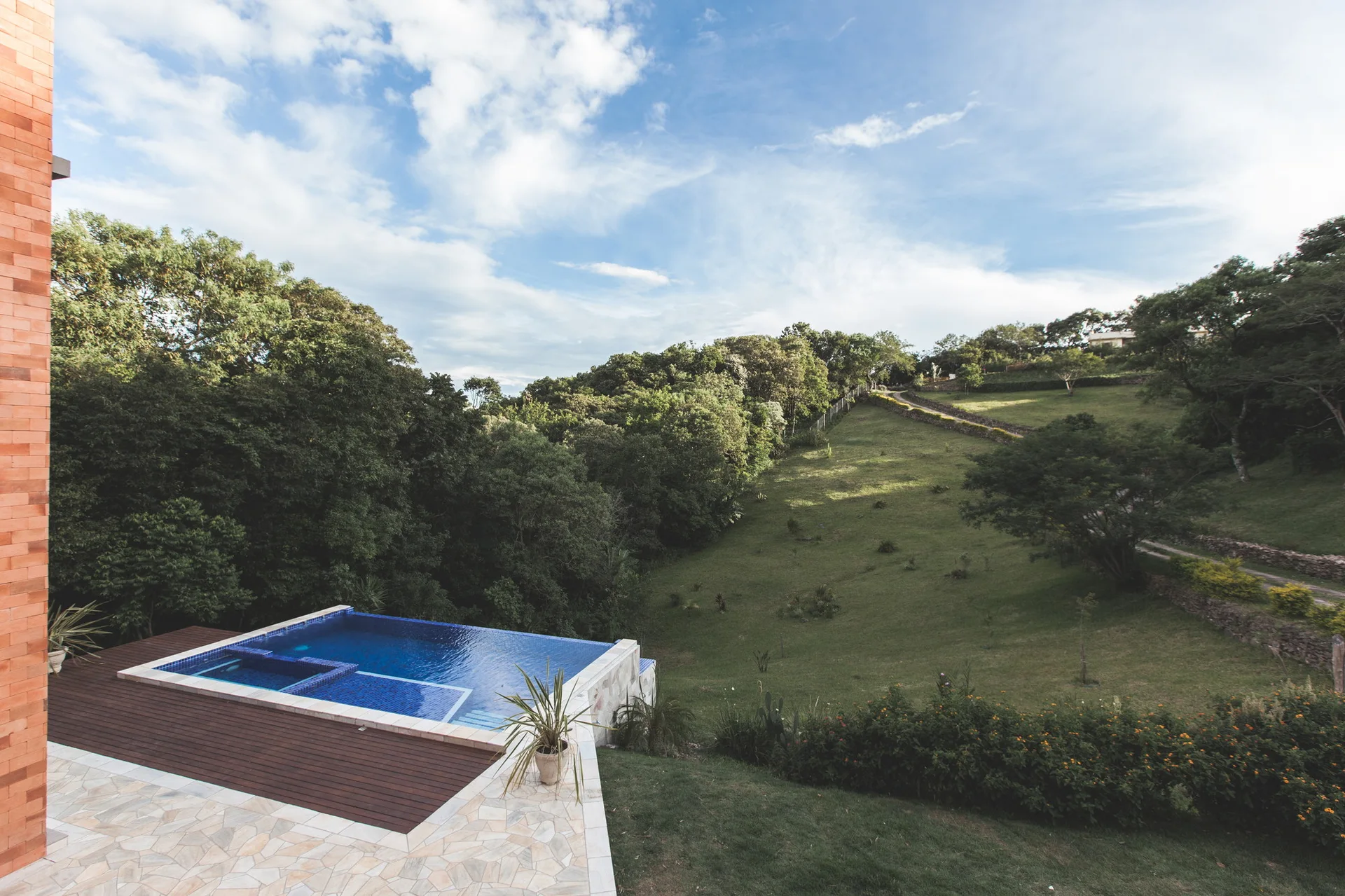 A blue tiled infinity pool on a hillside patio featuring dark wood decking, irregular stone pavers, and a red brick wall.