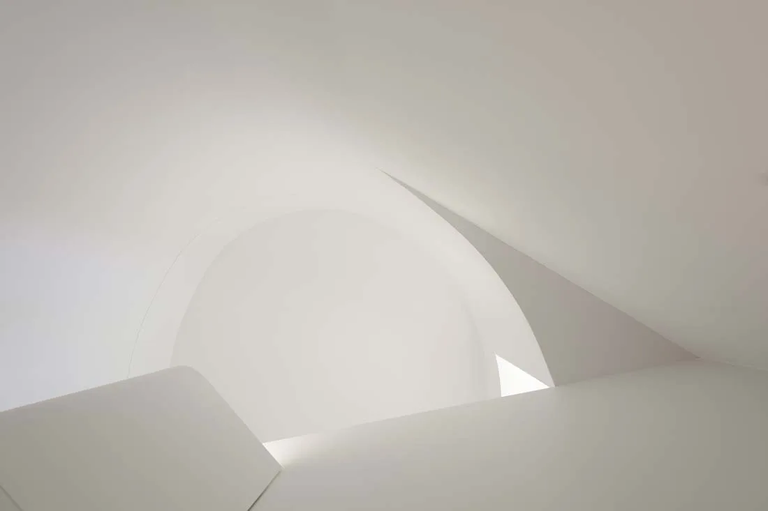 An upward view of a minimalist white interior featuring curved vaulted ceilings and clean architectural archways illuminated by soft light.