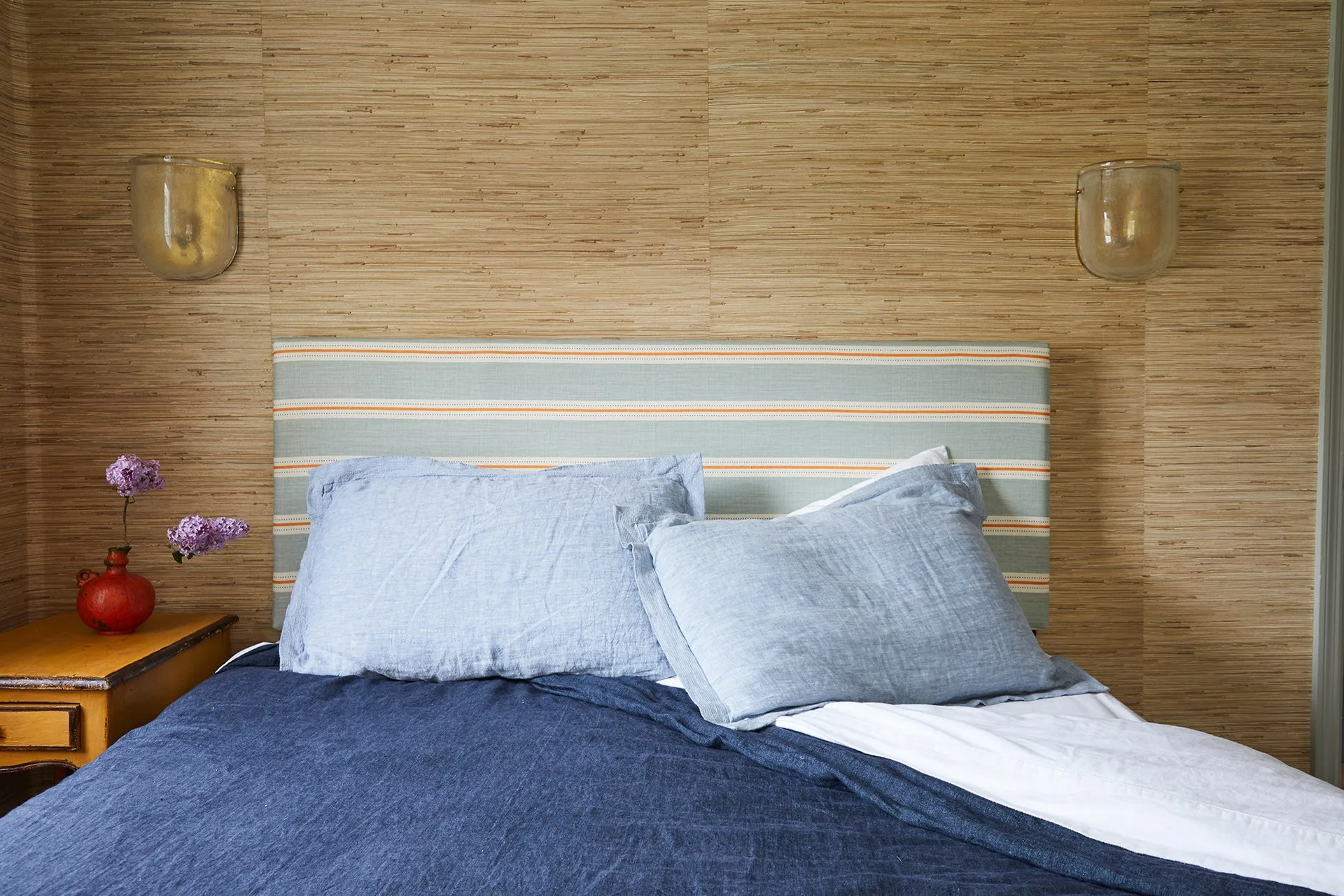 A bed with blue linen bedding and a striped headboard is set against a tan grasscloth wallpapered wall flanked by glass sconces.