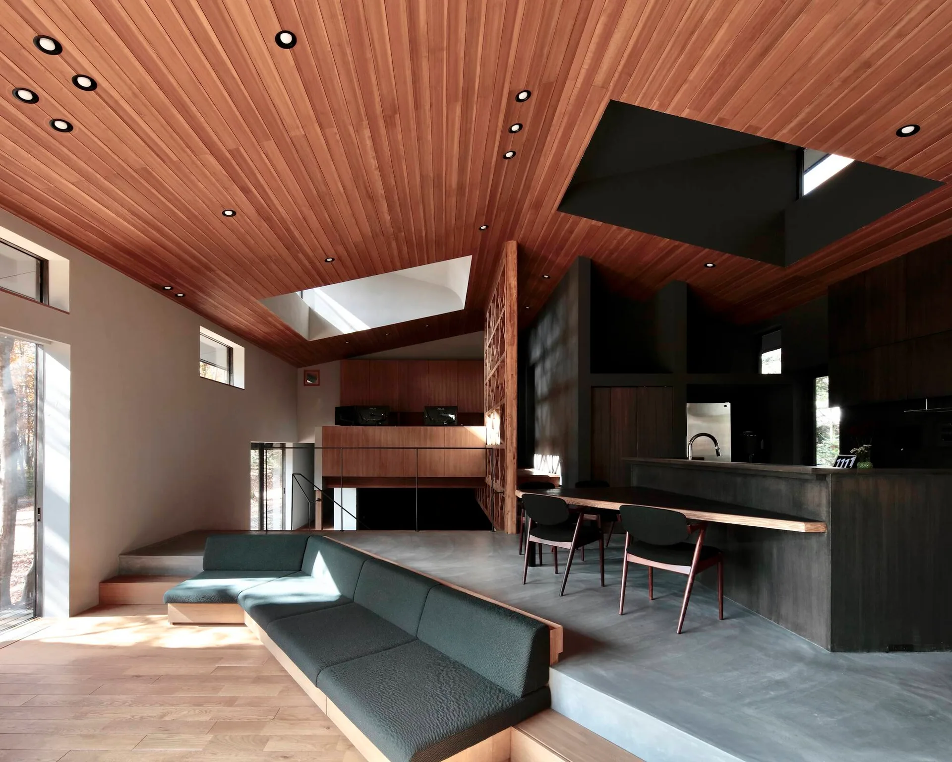 A modern open-plan interior with a slatted wood ceiling, a sunken grey sectional sofa, and a kitchen with dark cabinetry.