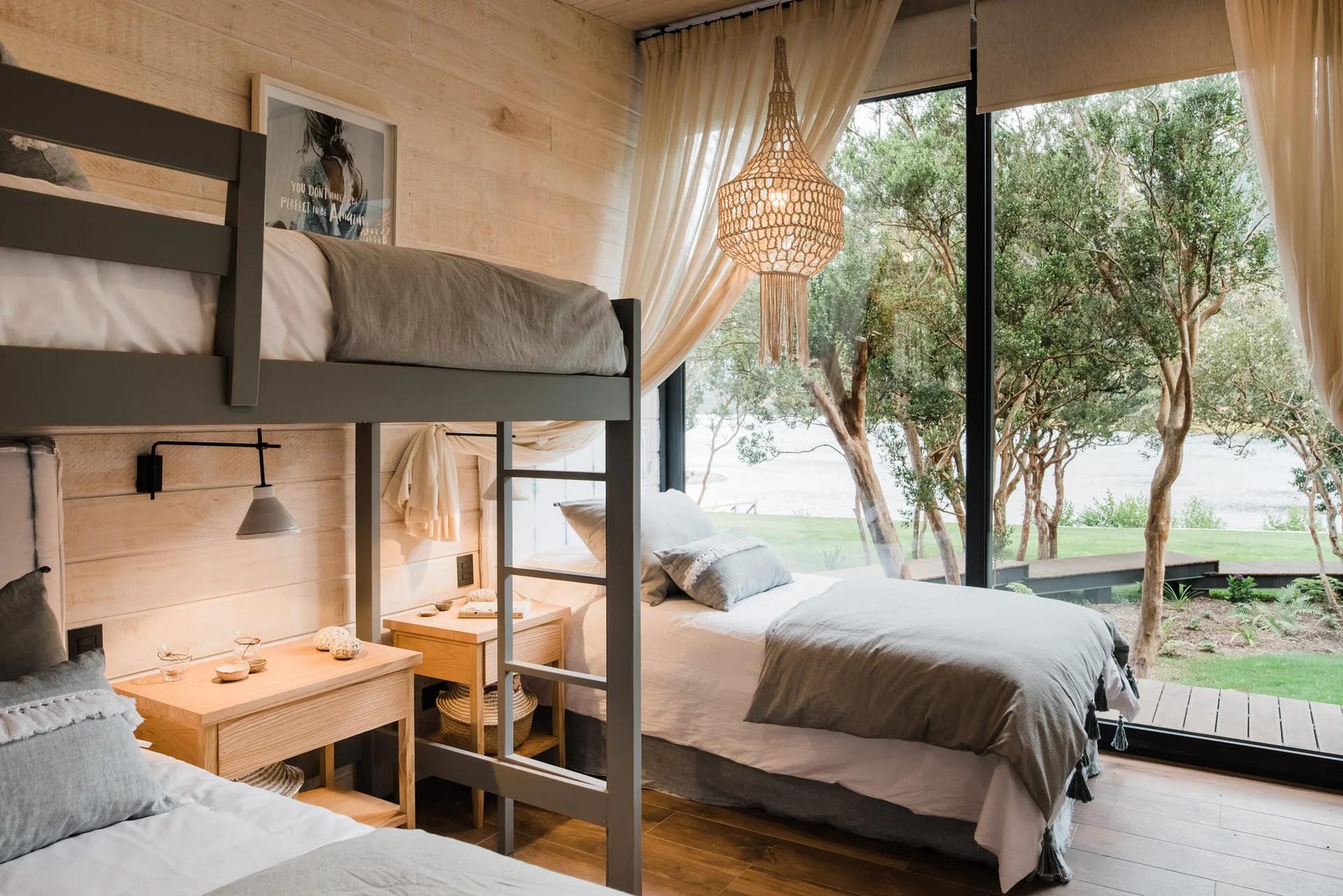 A light wood paneled bedroom features a gray bunk bed, a single bed by a floor-to-ceiling window, and a woven hanging light fixture.