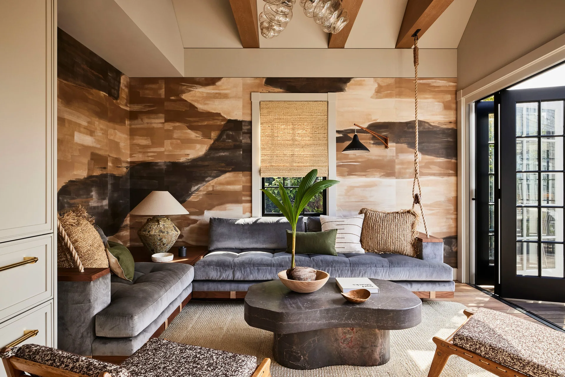 An organic modern living room features a grey velvet sectional, a sculptural stone coffee table, and an abstract brushstroke mural on the wall.
