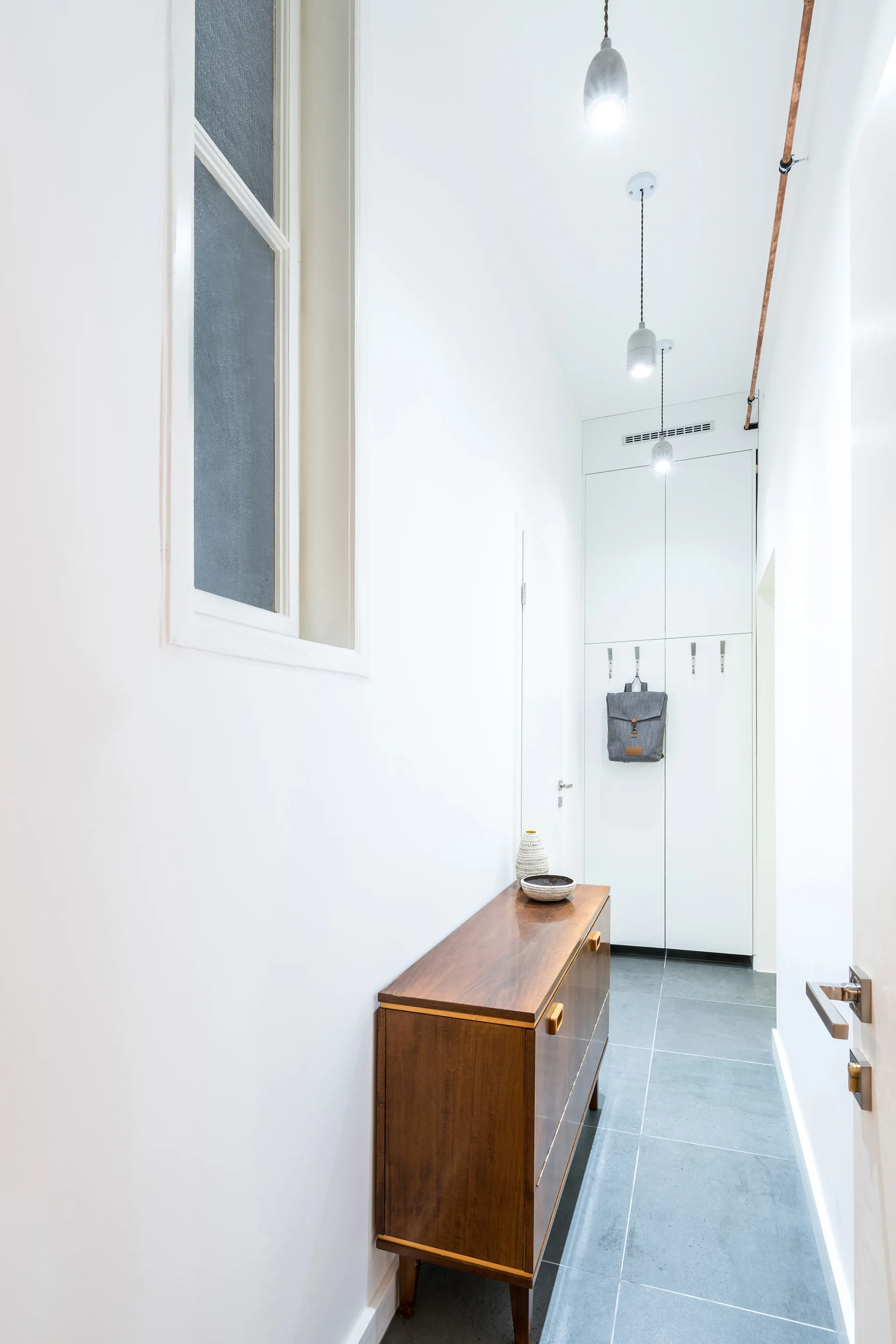 A long white hallway features a dark wood sideboard beneath a tall narrow window, grey floor tiles, and tall built-in storage cabinets.