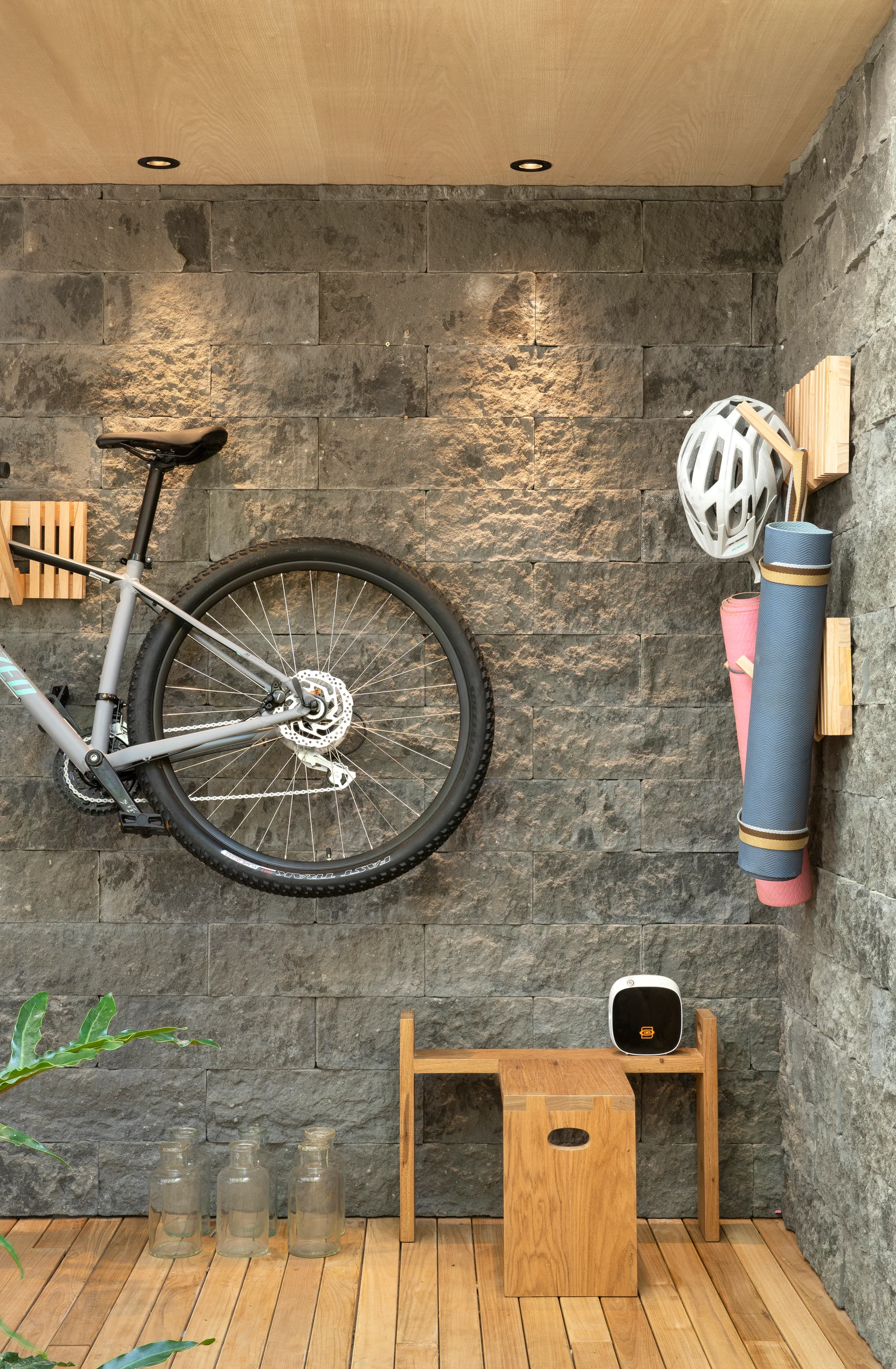 A grey mountain bike, helmet, and rolled yoga mats are mounted on a dark textured stone wall with wooden wall hooks above wood plank flooring.