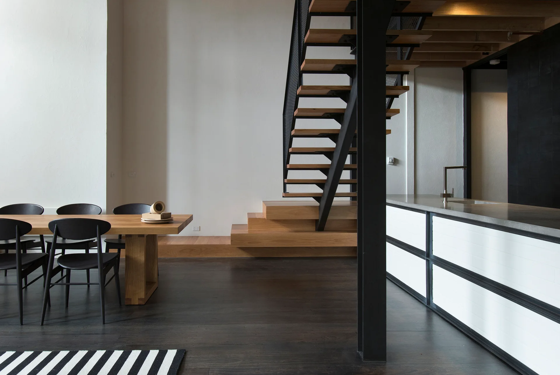 An open-plan dining space and kitchen featuring a wooden table with black chairs, a white kitchen island, and a black steel staircase with wood treads.